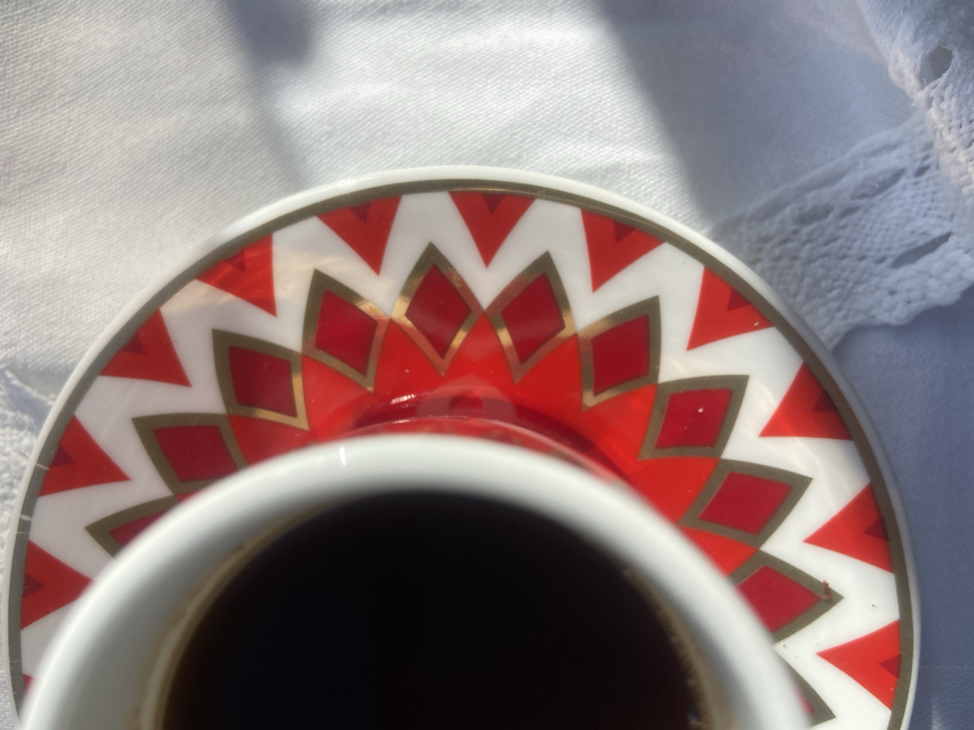 The image presents an intimate, top-down view of a white ceramic coffee cup and its matching saucer, resting on a white textured fabric. The cup, filled with a dark liquid (likely coffee), occupies the lower portion of the frame, with only its rim and the liquid contents visible.
The saucer is the focal point, adorned with a striking and colorful pattern. It features an inner ring of bright red diamonds, each meticulously outlined in gold. This is surrounded by an outer band where red and white triangles alternate, also delineated by fine gold lines, creating a geometric, zigzag effect. The gold accents catch the light, adding a subtle shimmer to the design.
The background consists of a soft, white fabric, possibly a tablecloth or linen, showing a slight texture and gentle folds. In the upper right corner, a delicate white lace trim is visible along the edge of the fabric, hinting at an elegant setting.
The scene is bathed in bright, natural sunlight, which casts distinct shadows across the white fabric in the upper right, indicating a clear, sunny day. The strong, directional light suggests either morning or late afternoon. The overall impression is one of a peaceful and enjoyable moment, perhaps a coffee break. No text is visible in the image. Although the location context is Budva, Montenegro, there are no specific visual cues within the frame to identify this location.
