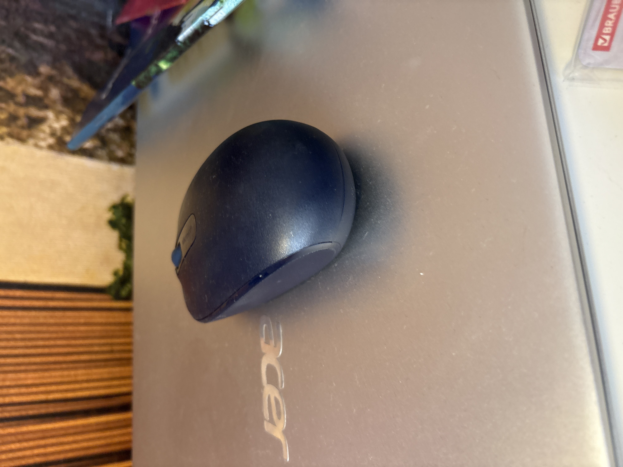 A dark blue computer mouse rests on the silver surface of a laptop with an "acer" logo visible. The background shows a textured wall with vertical lines and green foliage. The scene appears to be indoors, possibly on a desk or table. There is no indication of the location being in Georgiyevsk, Russia.