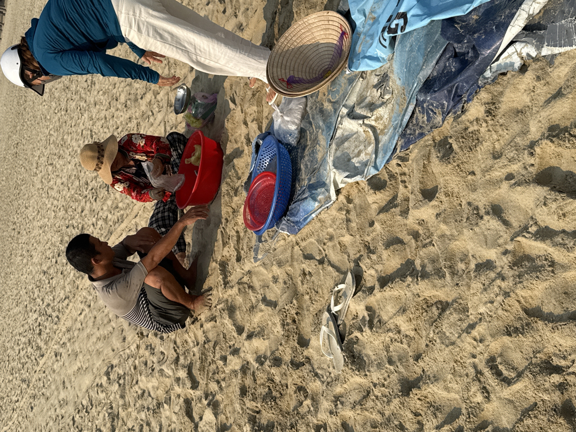 The scene is on a sandy beach in Đà Nẵng, Vietnam. Three people are present. One individual, wearing a white helmet, blue long-sleeved shirt, and white pants, is standing and reaching down towards a seated person wearing a floral shirt and a straw hat. This person is interacting with something in a red plastic tub. Adjacent to them, a man wearing a gray shirt and striped shorts is sitting on the sand.
Nearby objects include a conical straw hat, a blue tarp, blue and red plastic baskets, and a pair of flip-flops lying on the sand. The environment is sunny, casting shadows on the sand. The ground is sand.