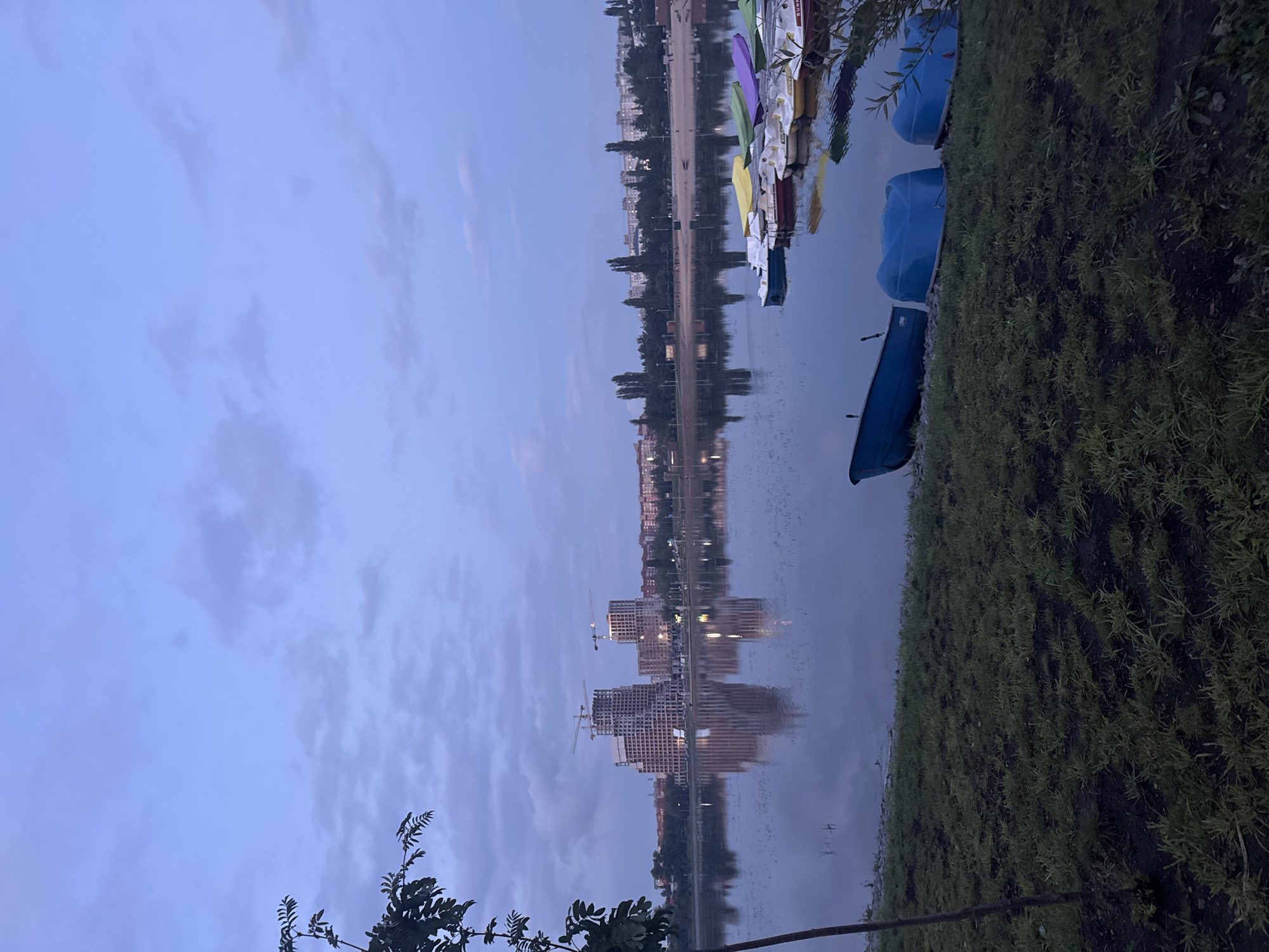 The image depicts a tranquil evening scene at a body of water, likely a river or lake, in Ufa, Russia. In the background, a city skyline is visible, characterized by several residential buildings and two prominent construction sites with cranes, suggesting ongoing development. The water's surface reflects the buildings and the dusky sky, creating a serene atmosphere.
In the foreground, a grassy bank slopes down to the water's edge. Several colorful paddle boats and smaller rowboats are moored along the shore. Some of these boats are partially on land, while others float gently on the water. A lone sapling with leafy branches stands on the left side of the frame. The sky is a muted blue with scattered, soft clouds, indicative of twilight or early evening. No people are actively engaged in activities within the frame, contributing to the peaceful ambiance. The overall impression is one of urban tranquility and the transition from day to night.