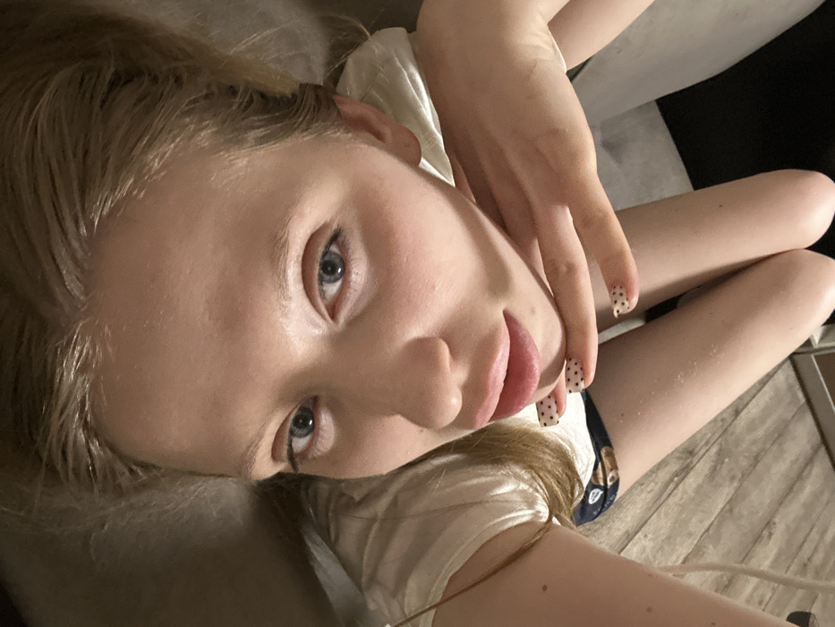 A young woman with fair skin, light blonde hair, and blue eyes gazes at the camera. She wears a white t-shirt. Her right hand rests near her face, displaying nails adorned with black polka dots. The image is slightly rotated, with the woman appearing to lie down or recline. The background features soft, dark hues and indistinct shapes.