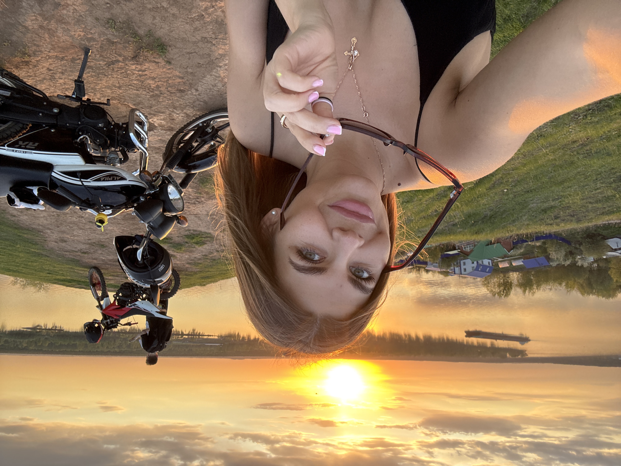 The image is an upside-down selfie of a woman. She is holding a pair of sunglasses. The woman is wearing a black top and a gold necklace with a cross pendant. A black and white motorcycle is visible in the background on the left side of the image. Further back, a man is next to a second motorcycle. The background consists of a landscape with water, sky, and a sunset. There are buildings on the far shore. Given the prompt, this is likely a location in Ольховка, Russia.