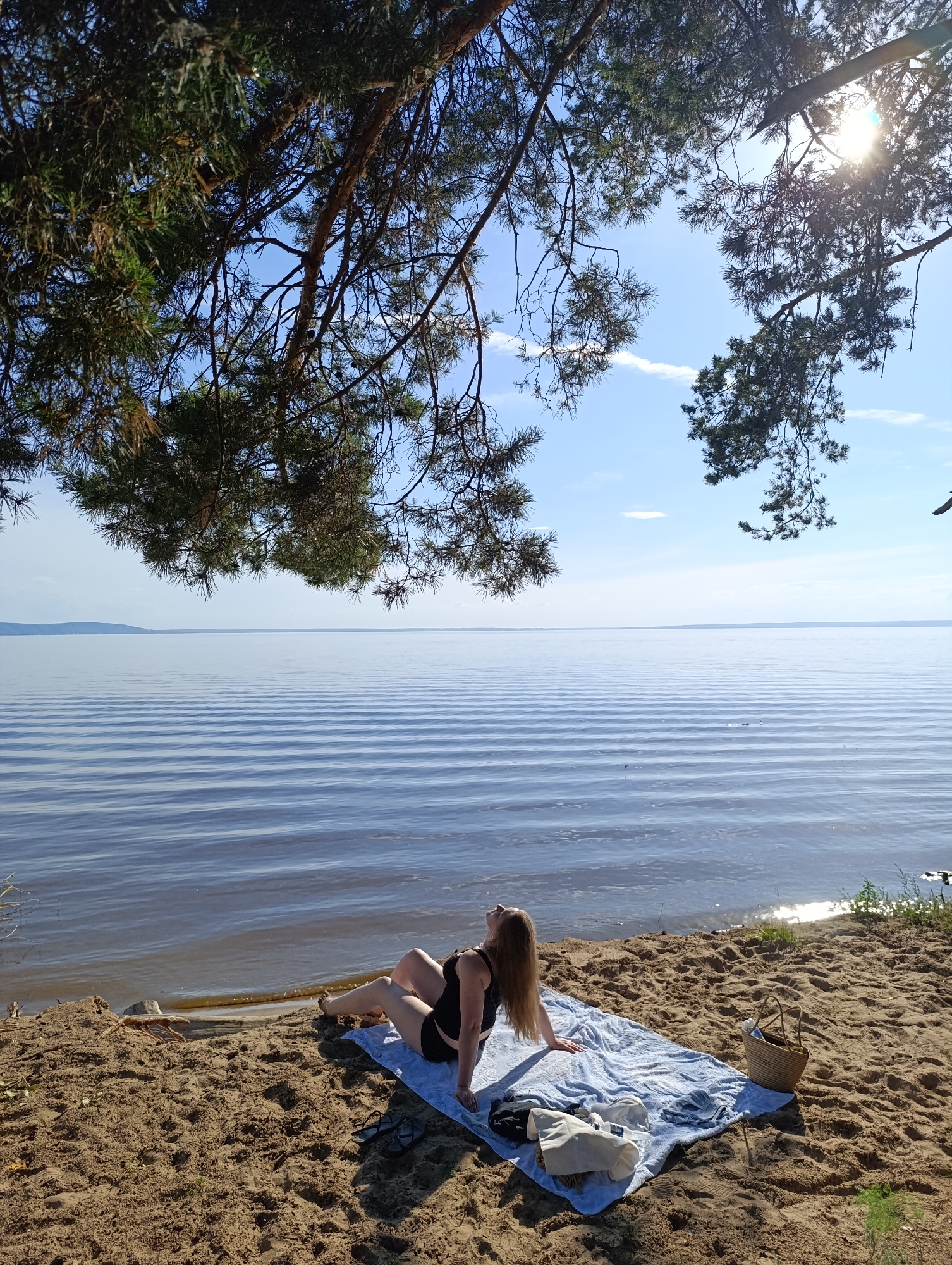 A woman is sunbathing on a beach next to a large body of water. She is wearing a black swimsuit and is reclining on a light blue blanket. There are several items on the blanket, including a straw bag, a white tote bag, and a pair of black sandals. The beach is made of sand and there are pine trees overhead, casting shadows on the scene. The water is calm with gentle ripples and reflects the bright sunlight. In the distance, a faint shoreline can be seen under a clear blue sky with a few wispy clouds. The sun is high in the sky, suggesting it is midday. The overall mood is peaceful and relaxed.
