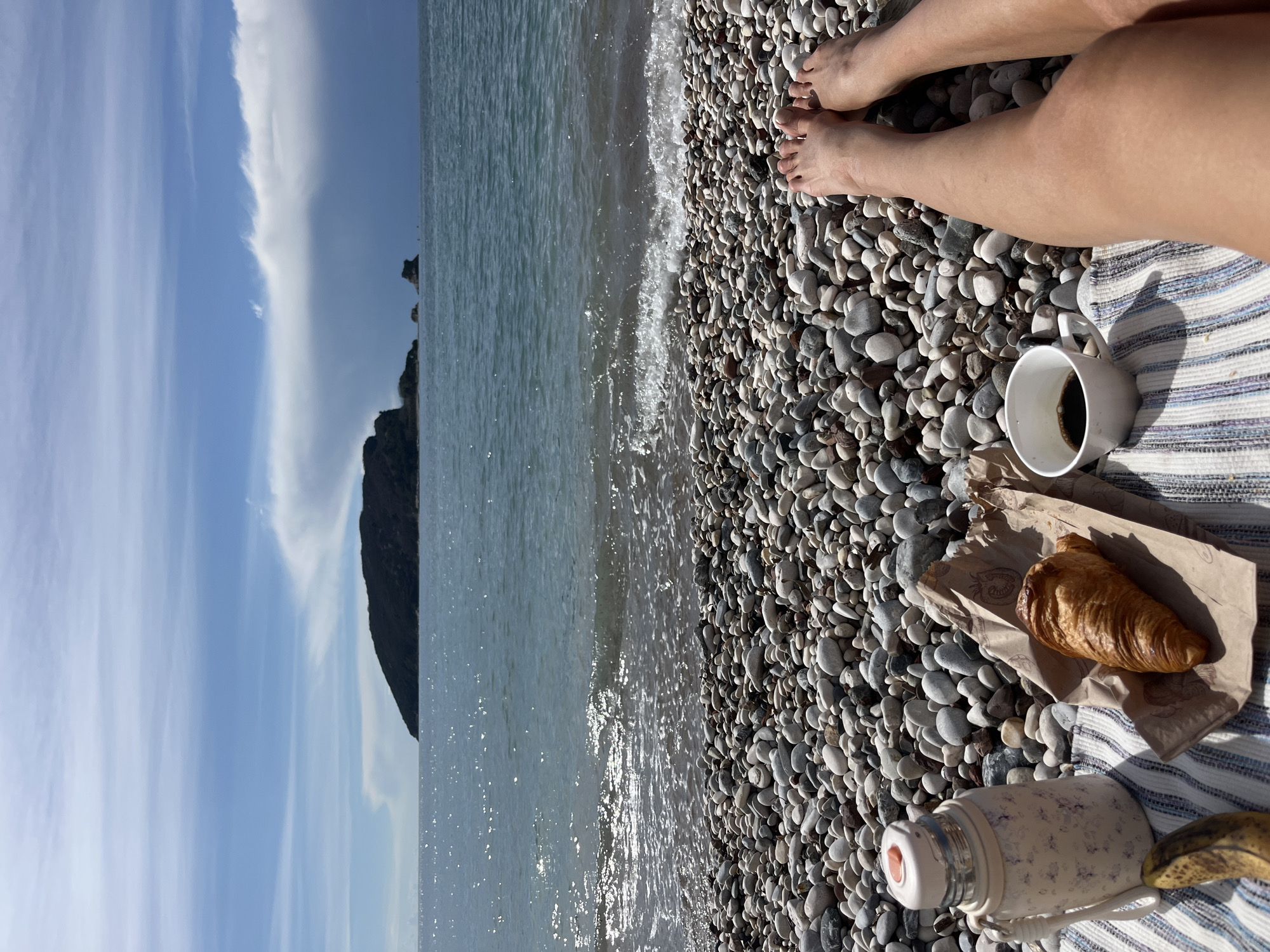 The image depicts a first-person perspective of a person relaxing on a pebble beach in Budva, Montenegro, with Sveti Nikola Island visible in the background.
In the foreground, the bare legs and feet of a person are visible, extended onto a multi-colored striped blanket resting on a bed of various-sized light grey and white pebbles. To the left, a cream-colored thermos with a floral pattern and a light orange cap stands upright. Next to it, a ripe banana with brown spots lies on the blanket. A croissant, golden-brown and flaky, rests on a piece of brown paper packaging with a faint strawberry logo. To the right, a white ceramic mug containing dark liquid, likely coffee, is positioned on the blanket.
The mid-ground features the Adriatic Sea, with gentle waves breaking on the pebble shoreline. The water exhibits a greenish-blue hue and reflects sunlight in sparkling patches.
In the background, Sveti Nikola Island, a dark, tree-covered landmass, rises from the sea, dominating the left side of the horizon. To its right, several smaller rocky islets are partially visible. The sky above is a mix of light blue with scattered white clouds, including a prominent, elongated white cloud formation stretching across the upper right portion, casting a subtle shadow on the distant sea.