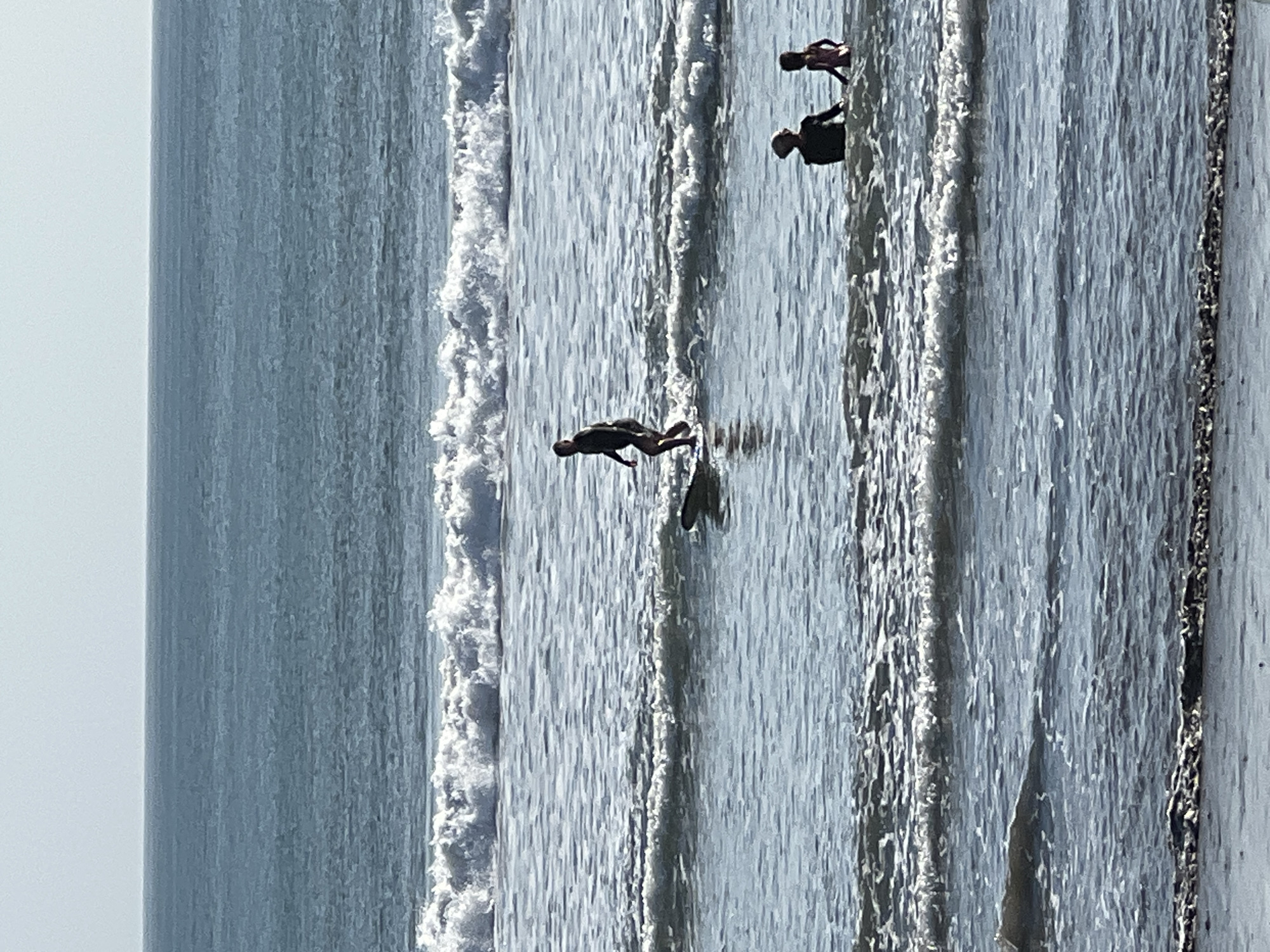 The image depicts a serene ocean scene, likely in Kuta, Indonesia, given the characteristic waves and tropical setting. The vast expanse of the ocean dominates the frame, with a clear, pale blue sky meeting the horizon. Gentle, foamy waves roll towards the shore, creating a rhythmic pattern across the water's surface. In the mid-ground, a lone surfer is riding a wave, their silhouette clearly defined against the lighter water. Further in the foreground, two individuals are wading in the shallow water, seemingly enjoying the ocean's embrace. The overall impression is one of peaceful recreation and the natural beauty of the sea.