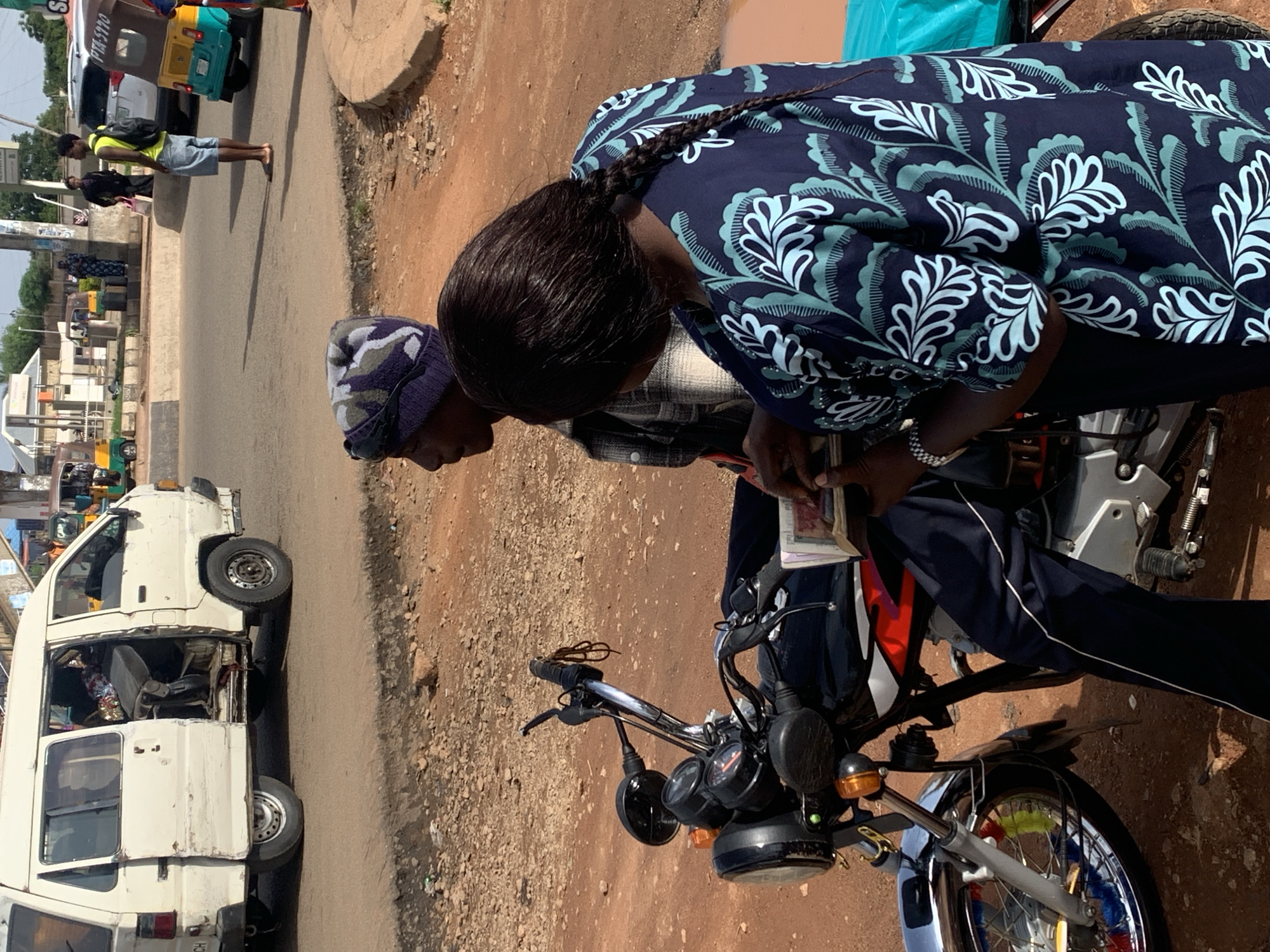 In Gura Top, Nigeria, a man wearing a blue and grey camouflage beanie and a plaid shirt is sitting on a motorcycle. A woman with dark hair in a braid, wearing a dark blue shirt with a white and teal leaf pattern, stands next to him, holding a stack of bills. The motorcycle is black with red and white accents. In the background, a white van with its door open is parked on a dirt road. Several other vehicles, including a colorful tuk-tuk, are visible further down the road, suggesting a busy street scene. The ground is predominantly red dirt and gravel, indicative of an unpaved area. The overall impression is a snapshot of daily life and commerce in a Nigerian town.
