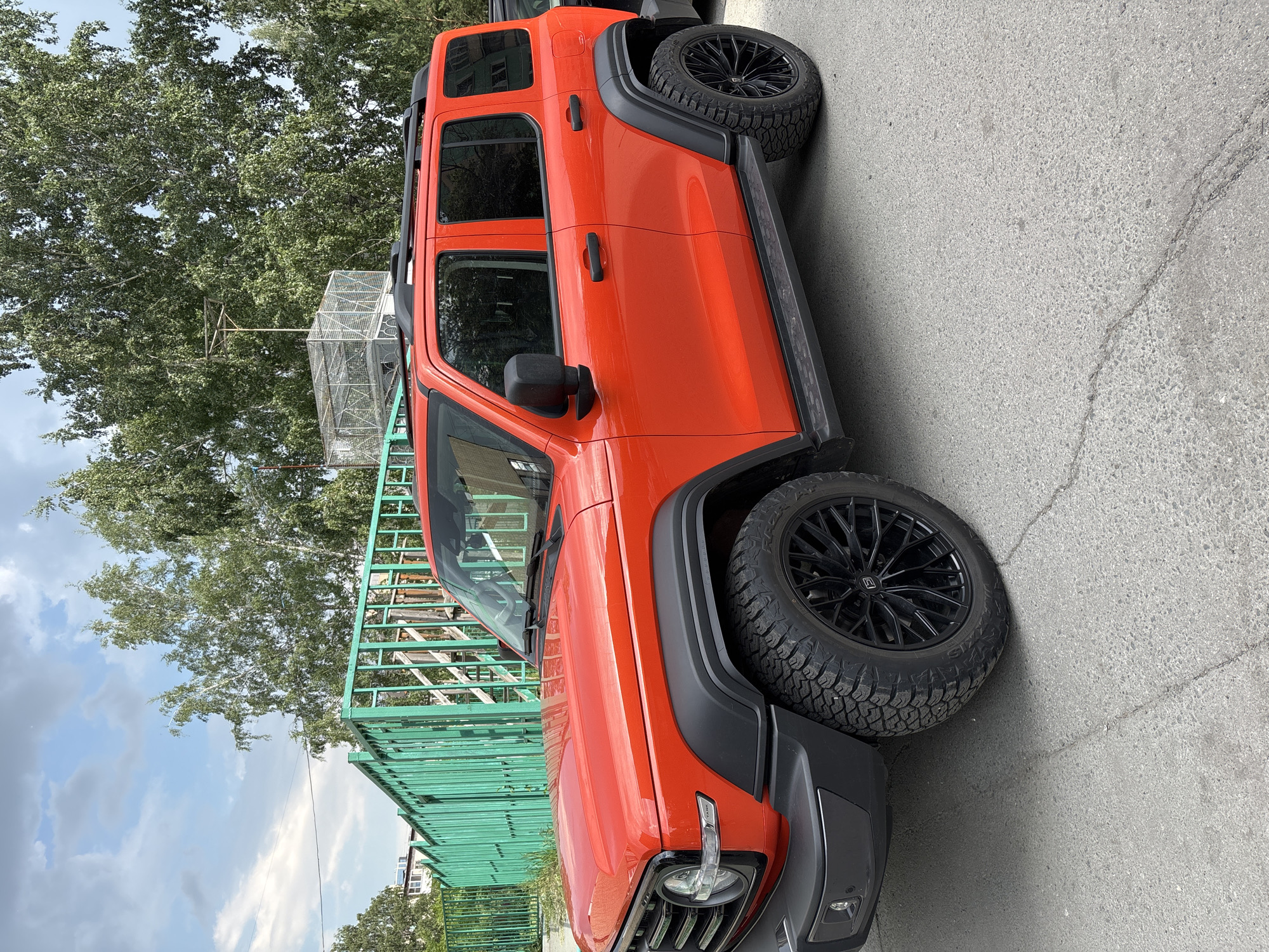 In Novosibirsk, Russia, a bright orange SUV, likely a TANK 300, is parked on a cracked asphalt surface. The vehicle features black fender flares, black multi-spoke wheels with aggressive tires, and a black roof rack. To the left, a vibrant green metal fence runs along the edge of a paved area. Behind the fence, trees with lush green foliage are visible, and further in the background, a multi-story building can be partially seen. The sky above is partly cloudy with a mix of blue and white. The scene appears to be an outdoor urban setting on a clear day.