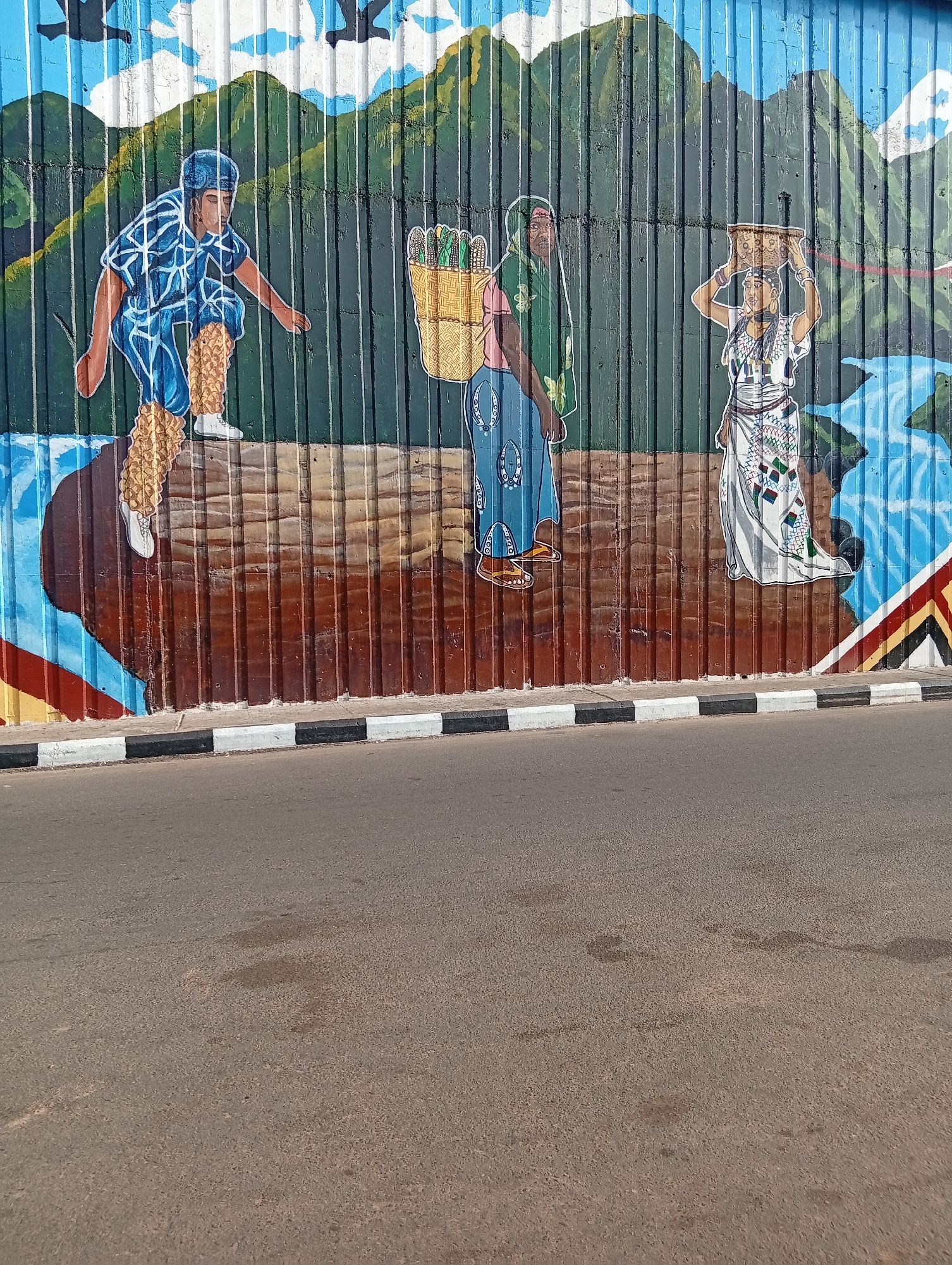 This is a mural painted on a corrugated metal wall, located in Jalingo, Nigeria. The mural depicts three people in traditional attire. On the left, a person in a blue and white patterned outfit, with what appears to be fur leg coverings, crouches. In the center, a person in a green headscarf and a blue dress carries a basket of what looks like corn. To the right, a person in a white patterned dress balances a woven basket on their head. Behind the figures, the mural shows a mountainous landscape with a blue sky and birds. A river is depicted flowing on the right side of the mural. The mural is situated above a street, with a curb featuring black and white stripes in the foreground. The road itself is paved and shows some wear and tear, with darker patches possibly indicating oil or water stains. The overall impression is one of cultural depiction and local artistry.