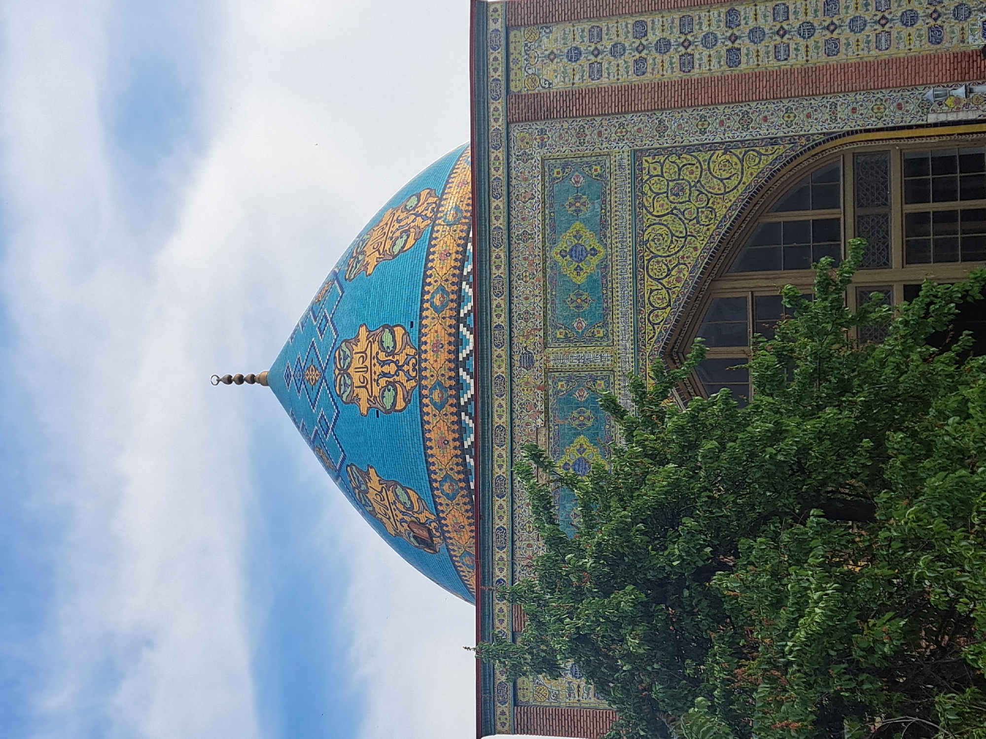 The image shows the Blue Mosque in Yerevan, Armenia. The architecture is predominantly Islamic, with a large blue tiled dome adorned with gold patterns. The adjacent wall is decorated with intricate geometric and floral designs in various colors, including blue, yellow, and turquoise. A large arched window is visible on the right side of the wall. In the foreground, a green tree partially obscures the view. The sky, visible behind the dome, is light blue with scattered clouds.