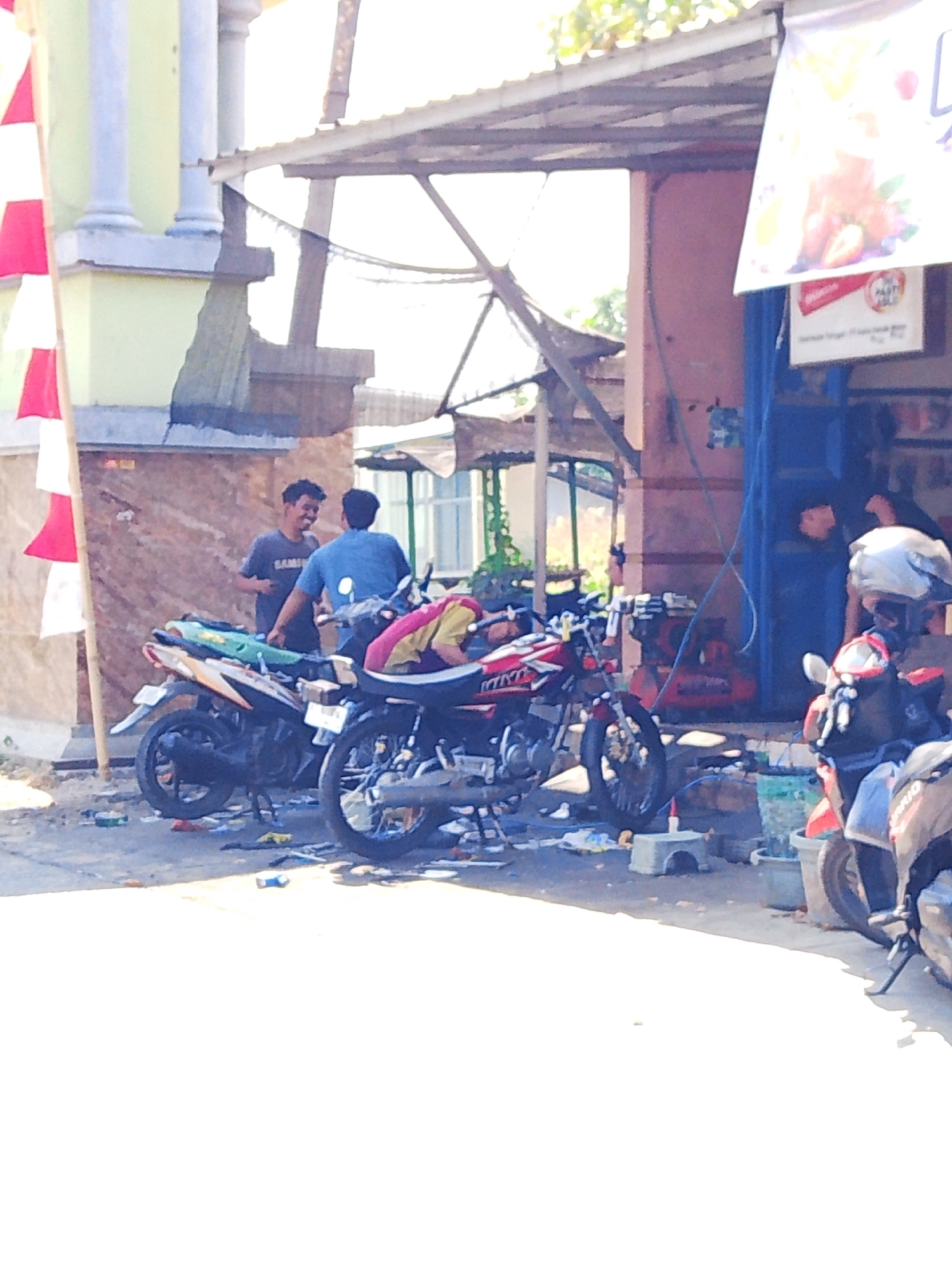 In the city of Kedondong, Indonesia, a street scene unfolds under a bright, possibly sunny sky. Two men are interacting near several parked motorcycles. One man, wearing a dark t-shirt with the word "SAM" visible on it, stands with his left arm extended, appearing to gesture or talk. He has dark, curly hair. The second man, dressed in a blue t-shirt, faces away from the camera and is engaged in conversation with the first man.
In the foreground and midground, multiple motorcycles are present. One prominent motorcycle in the center is red and black with yellow accents, and appears to be on a kickstand. Another motorcycle to the left is orange and black. A third motorcycle, mostly visible on the right, is red and black and has a white helmet on its handlebars. The ground around the motorcycles is littered with debris, including plastic wrappers and scattered objects.
Behind the men and motorcycles, a building structure with a corrugated metal roof is visible. To the left, a building with light green walls and light blue pillars stands. A red and white striped flag is attached to a pole near this building. Further back, a dark blue tarp or fabric is stretched taut. The overall environment appears to be an outdoor, possibly commercial or residential area, with a slightly unkempt appearance due to the scattered debris. The image quality is somewhat grainy and the focus is not sharp throughout.