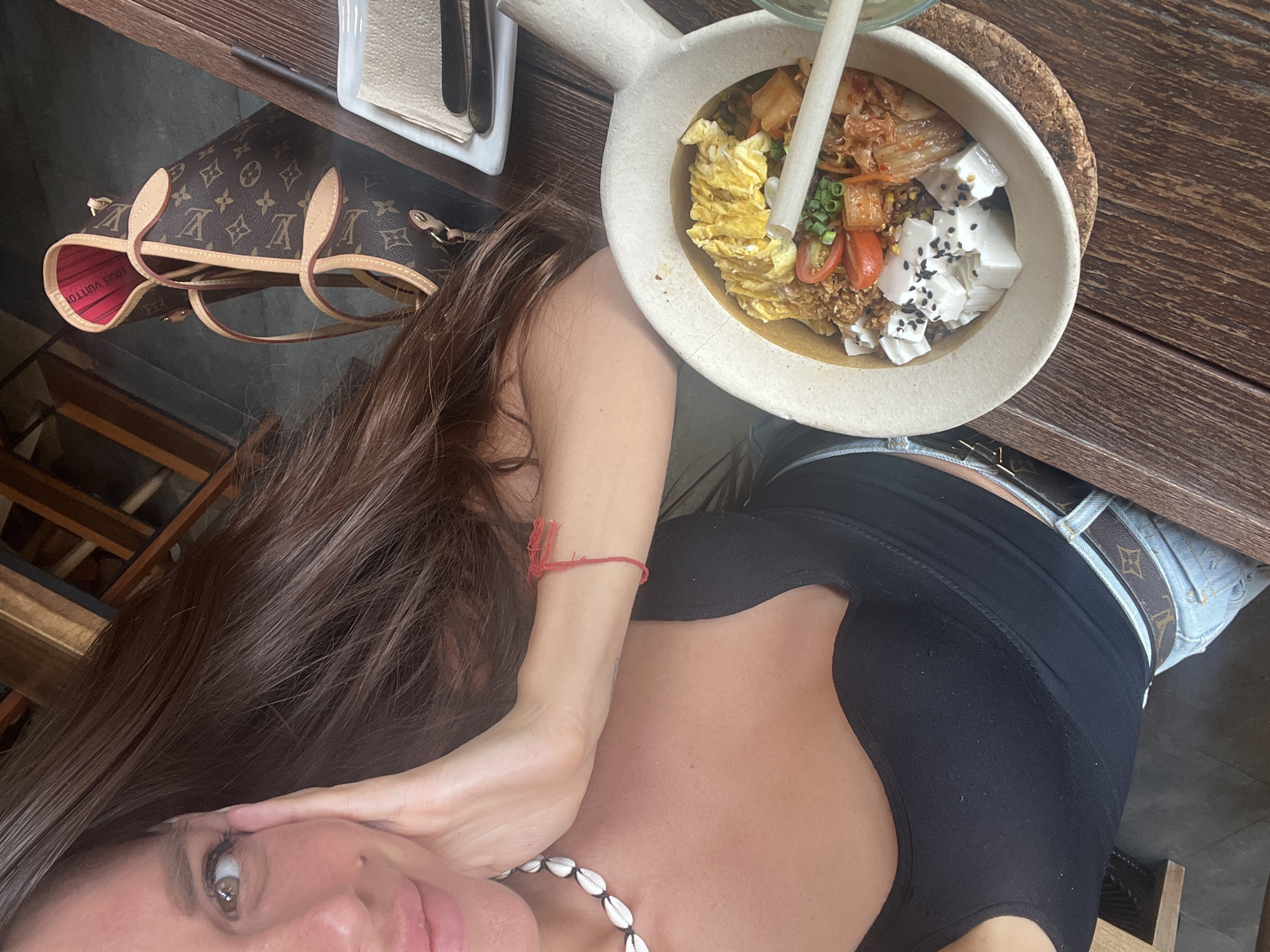 A woman with long brown hair and a black tank top is shown from the chest up, resting her hand on her cheek. She is wearing a necklace made of shells and a red string bracelet. A bowl of food containing rice, kimchi, tomatoes, tofu, and scrambled eggs is in the foreground, to the right of the woman. A Louis Vuitton purse is visible behind her. The setting appears to be a cafe or restaurant with a wooden table and floor.