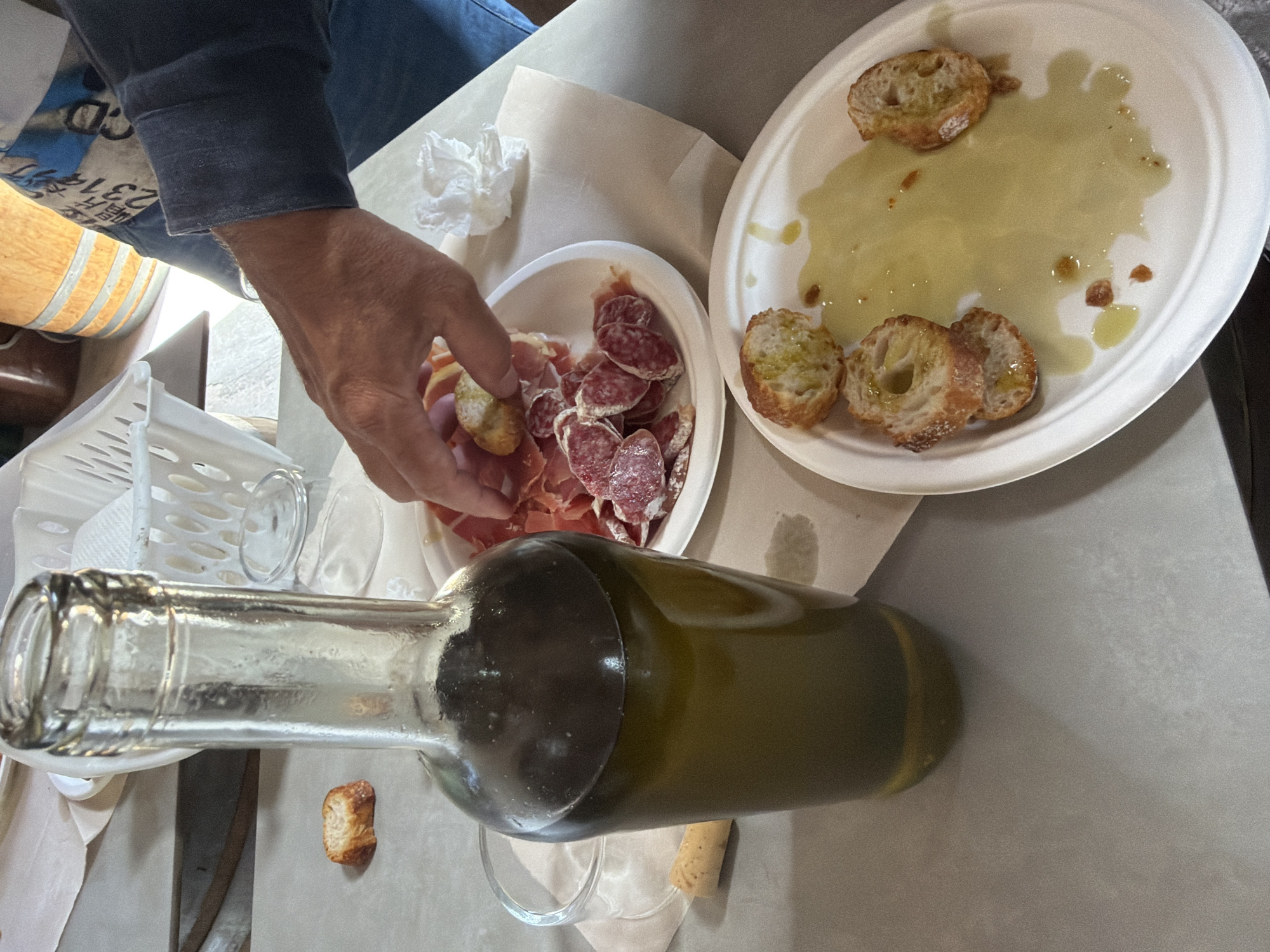 A top-down, slightly angled shot captures a casual food and drink setting, likely a tasting, in Bonastre, Spain. In the foreground, a dark green bottle, possibly olive oil or wine, lies horizontally across the table, with its neck extending towards the left and its base towards the right. An empty, clear glass and a cork are visible near the bottle's base.
A person's arm and hand are prominent on the left side, reaching towards a white, disposable plate filled with an assortment of cured meats, including thin slices of what appears to be prosciutto and round slices of salami. The hand is holding a piece of crusty bread. To the right of this plate is another similar disposable plate, largely consumed, featuring several pieces of bread soaked in a puddle of olive oil with a few darker flecks, suggesting herbs or spices. A crumpled white napkin rests on the table near the person's hand.
The table surface is covered with a light-colored material, possibly paper or a light cloth. In the blurred background, a wooden barrel with metal bands is partially visible, hinting at a winery or an olive oil farm. On the sleeve of the person's dark blue shirt, faint white text and numbers, such as "CD" and "2314", are discernible but not fully legible. The scene depicts an ongoing snack or tasting, characterized by simple, rustic fare.
