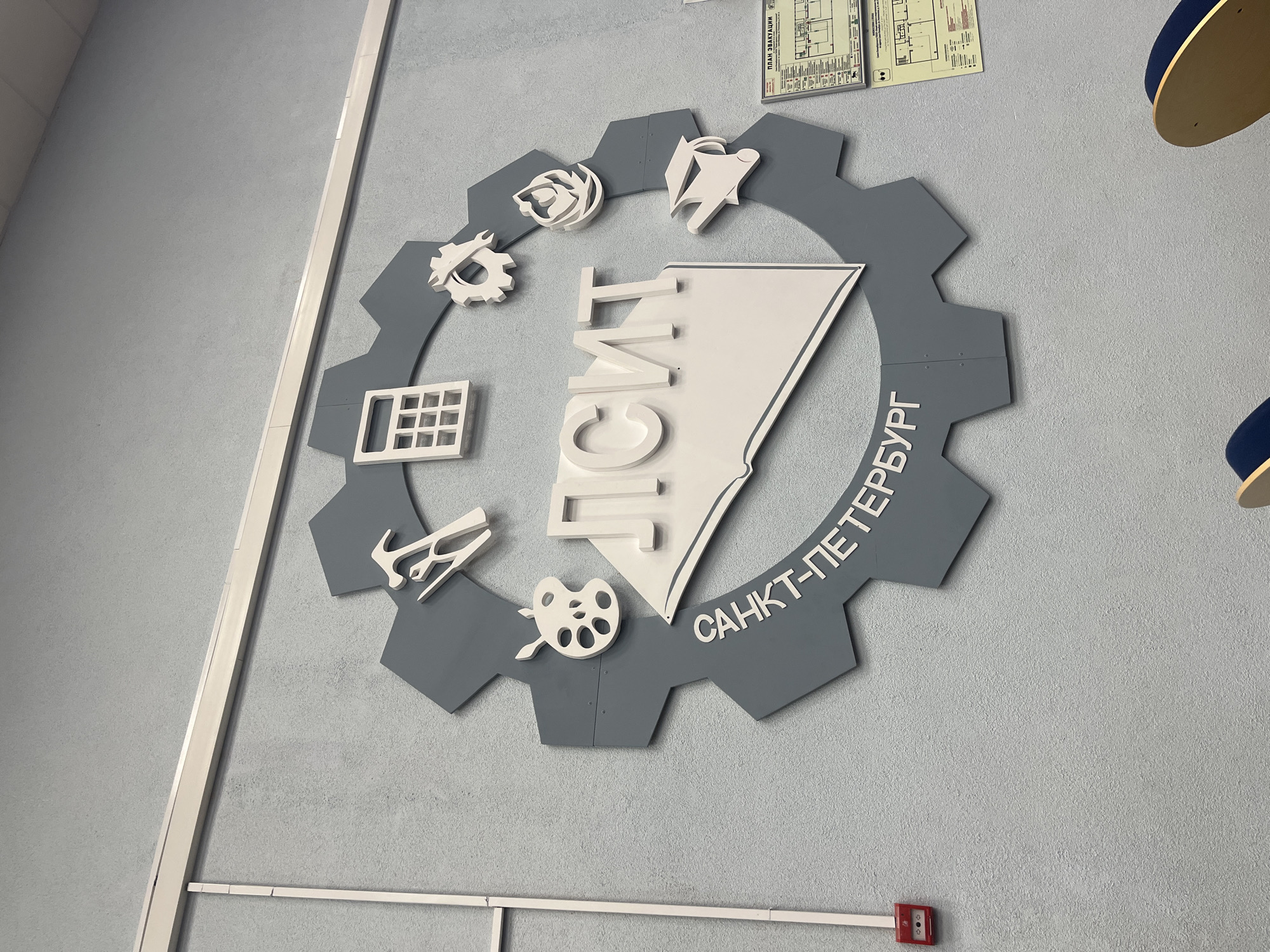 The image shows a sign on a light blue wall. The sign is shaped like a gray gear with a white triangular shape in the center, containing the vertically oriented word "СПБГУПТД" (Saint Petersburg State University of Industrial Technologies and Design). Around the gear are several white icons, including a calculator, tools, an artist's palette, a wrench, and others. The text "САНКТ-ПЕТЕРБУРГ" (Saint Petersburg) is written along the lower right side of the gear. There is a white trim on the left side of the wall. Near the bottom of the image are horizontal white bars with a red emergency fire alarm. The location is Saint Petersburg. The location in the city is Novoe Devyatkino, Russia.