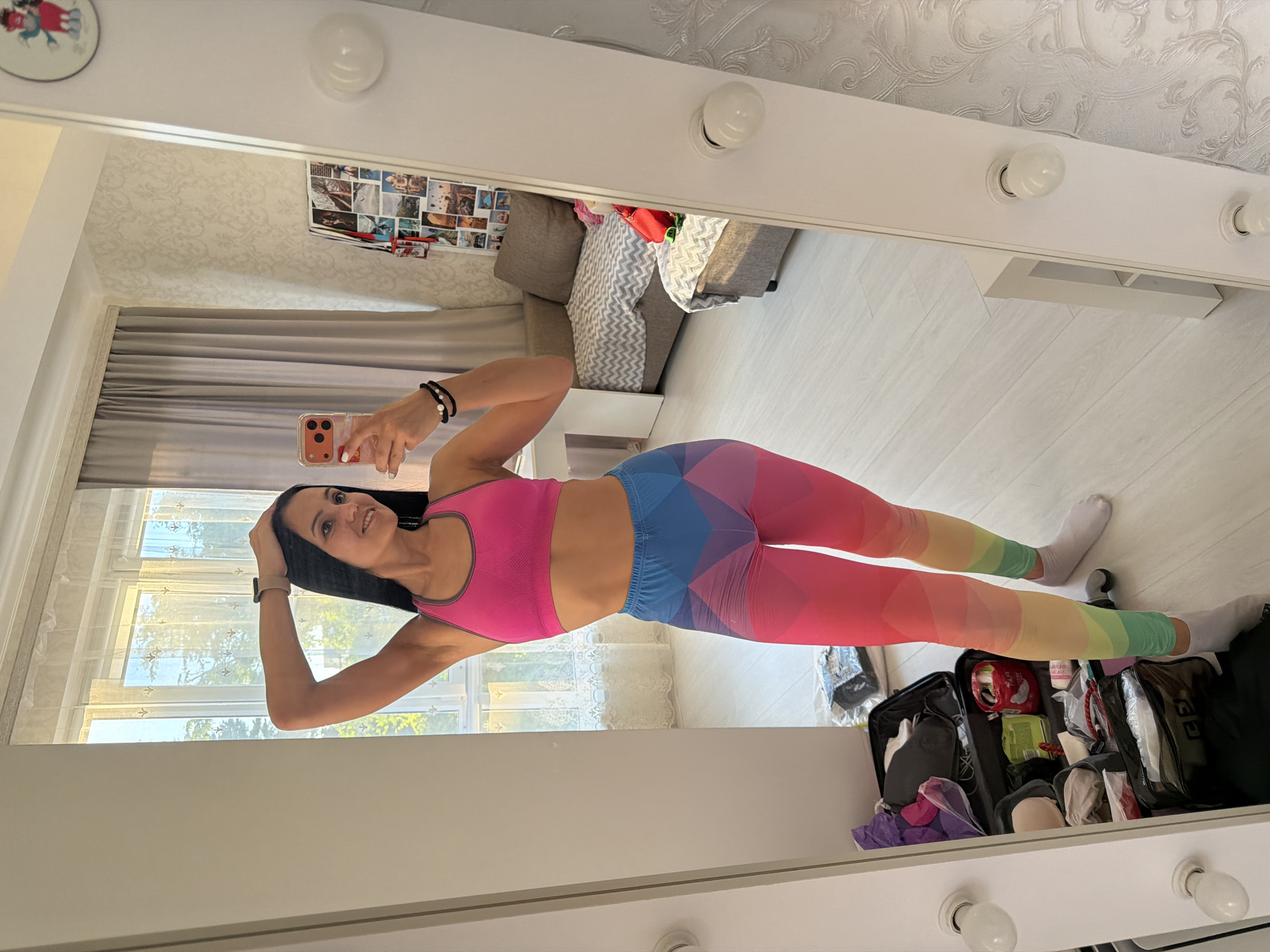 A woman is taking a selfie in a mirror. She has long dark hair and is wearing a pink sports bra and colorful leggings. The leggings have a geometric pattern that transitions through the colors of the rainbow from her waist to her ankles. She is standing in a room with white walls and light-colored wood flooring. In the background, a couch and a window with sheer curtains are visible. To the right of the woman, a black suitcase is open and filled with various items, including makeup bags, bottles, and clothing. The overall lighting suggests it is daytime. The woman's expression is cheerful and engaged. The location is identified as Sochi, Russia.