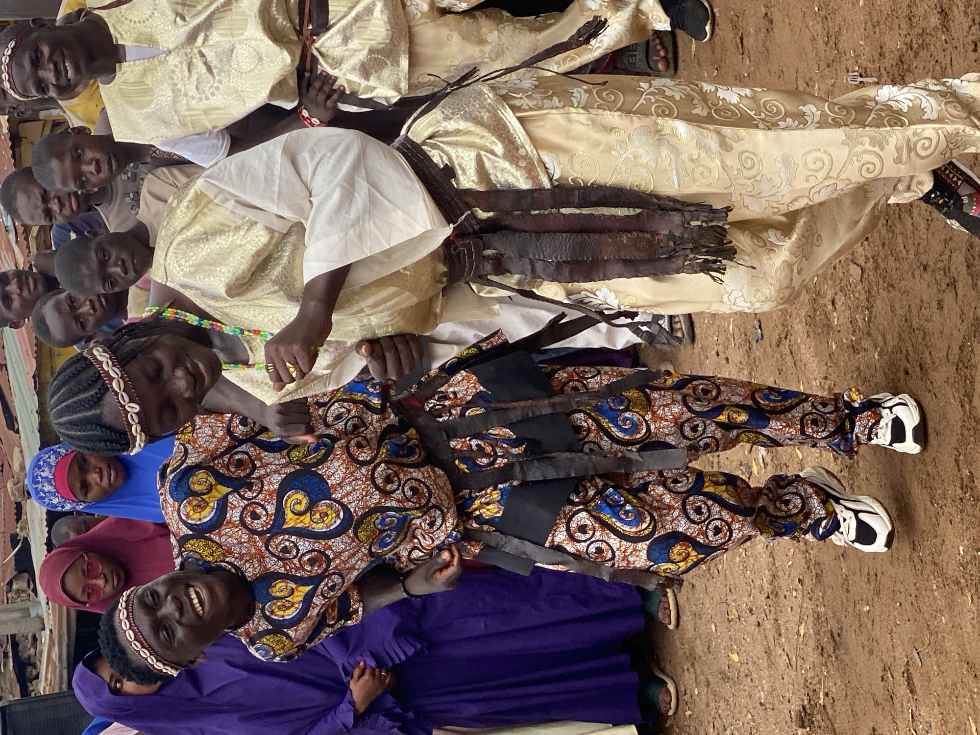 This media file depicts a group of people, predominantly children and young adults, gathered outdoors in what appears to be a rural or semi-rural setting in Rigachikun, Nigeria. The ground is dirt, suggesting an unpaved area. The individuals are dressed in a variety of clothing. Some are wearing traditional attire, including ornate, possibly ceremonial, garments in cream and gold fabric with intricate patterns. Others are in more casual clothing, such as t-shirts and head coverings (hijabs) in colors like blue and burgundy.
One prominent subject, a young woman or girl, is in the foreground, wearing a vibrant, patterned outfit with a colorful print featuring swirls of blue, yellow, orange, and black. She has dark braided hair adorned with a band of small white shells and is smiling broadly, her teeth visible. She is also wearing a beaded necklace in multi-colors. Another individual, possibly an elder, is partially visible in the background, dressed in a similar ornate gold garment.
Several younger individuals are also visible, some looking towards the camera with curious or smiling expressions. The overall mood appears to be lively and communal, possibly during a local gathering, celebration, or event. The presence of multiple individuals, some in celebratory attire, suggests a special occasion. The lighting suggests it is daytime, likely under natural sunlight, though the specific time of day is not clear. There is no visible text in the image.