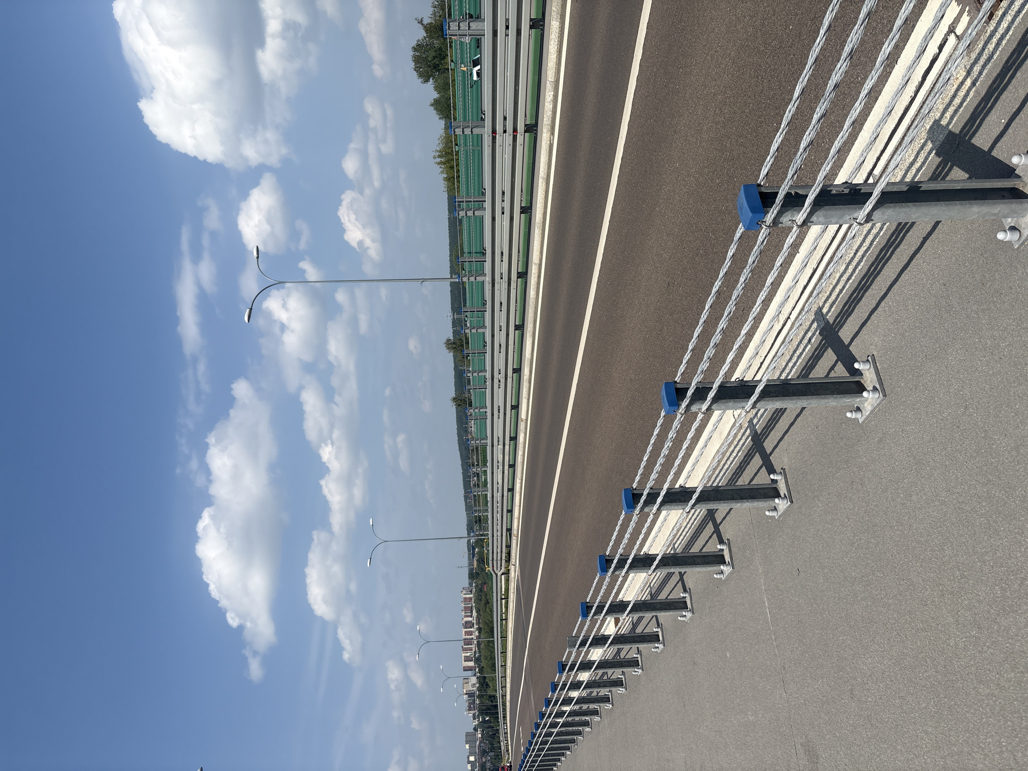 An outdoor, eye-level, medium shot shows a bridge on a sunny day. The sky is blue with scattered white clouds. In the foreground, a grey asphalt sidewalk runs along the left side of the image, separated from the road by a metal railing. The railing consists of vertical metal posts with horizontal steel cables strung between them, topped with blue caps. The road, also made of dark asphalt, runs parallel to the sidewalk and is divided by a white line. Beyond the road, a green sound barrier is visible, with horizontal metal railings in front of it. Streetlights with curved arms are spaced along the roadside. In the background, a city skyline with buildings and trees can be seen under the bright sky.