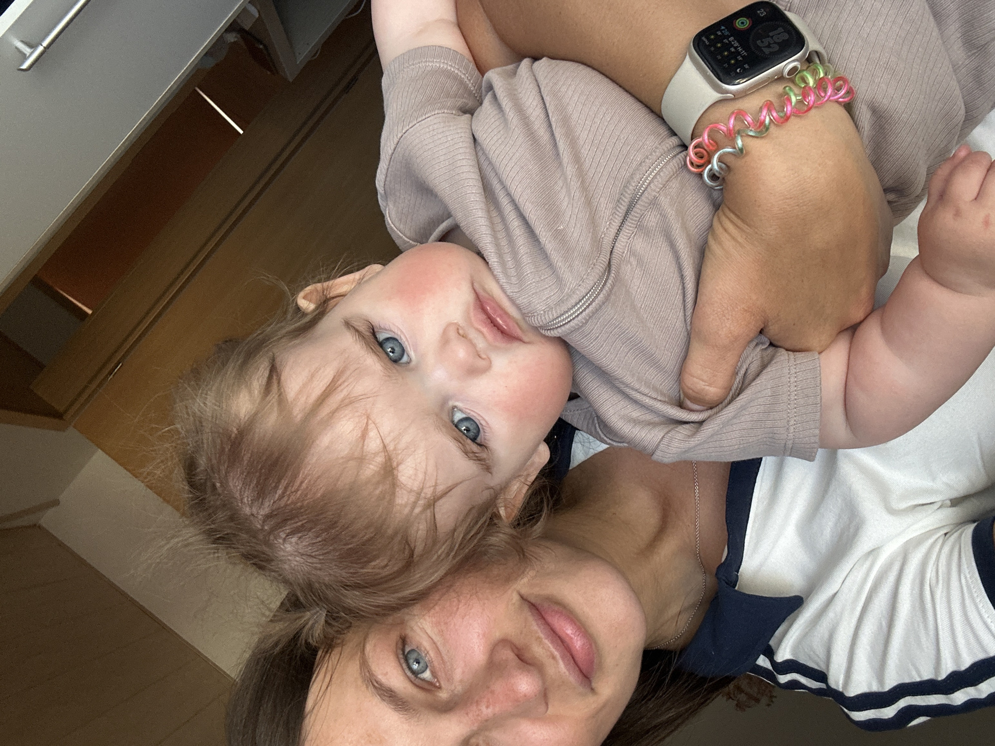 A woman and a young child are featured in a close-up, indoor selfie taken from a slightly upward angle. The child, dressed in a light brown ribbed jacket, is held in the woman's arms. The woman is wearing a white shirt with navy blue trim and a silver chain necklace. An Apple watch is visible on her wrist, accompanied by a colorful spiral bracelet. The background shows part of a cabinet or storage unit, suggesting a domestic setting. There is no available evidence to indicate that the location is in Niederquembach, Germany.
