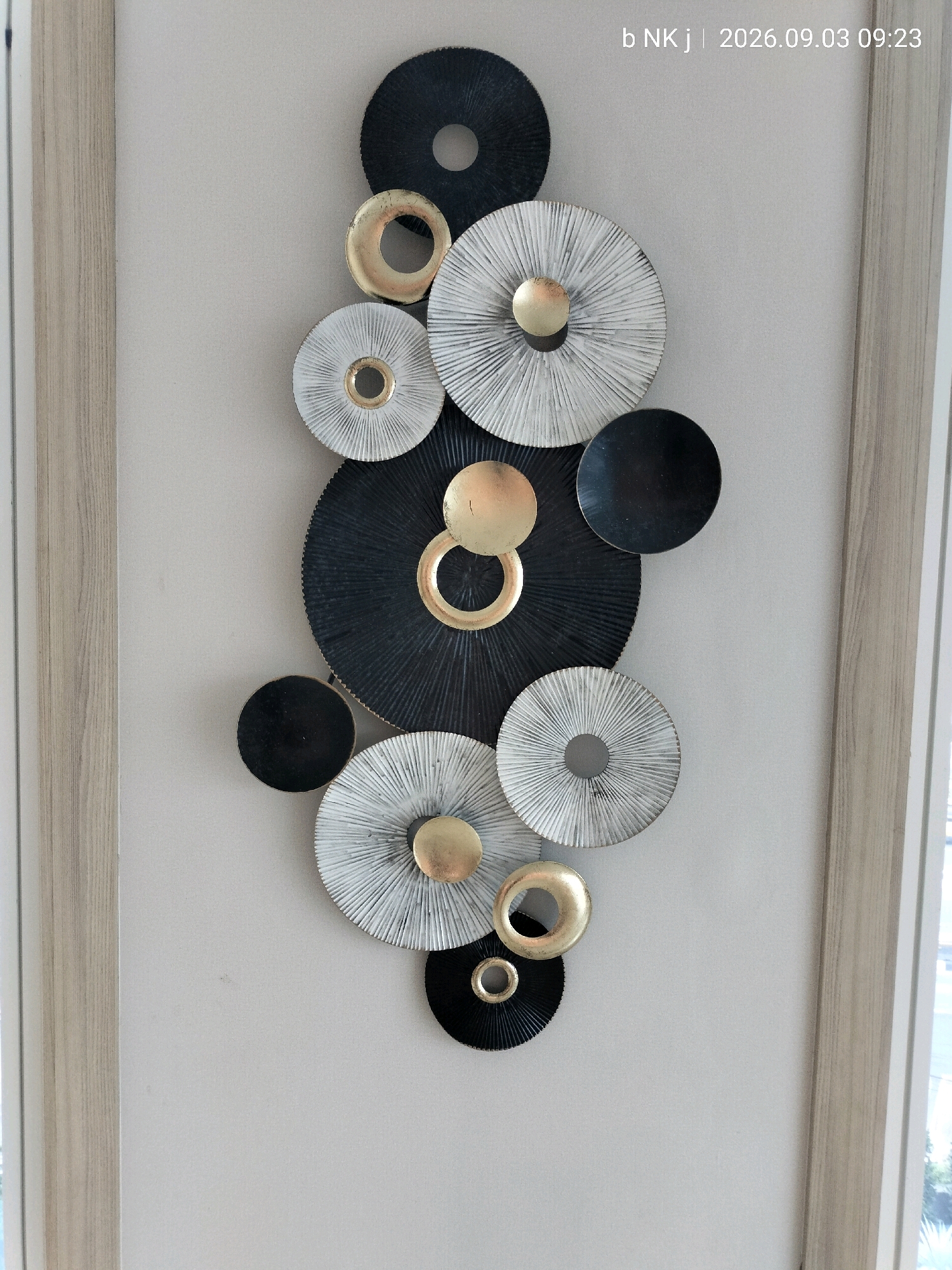 The image shows a wall decoration composed of several circular metal pieces in varying sizes and colors, including black, white with textured radiating lines, and gold. The arrangement is asymmetrical and vertical, with the largest black piece near the center and smaller pieces surrounding it. The wall itself is a light, neutral color, and a wooden trim or door frame is visible on the left side of the image. A timestamp and text are superimposed on the upper right corner of the image, reading "b NK j | 2026.09.03 09:23". The location is South Jakarta, Indonesia.