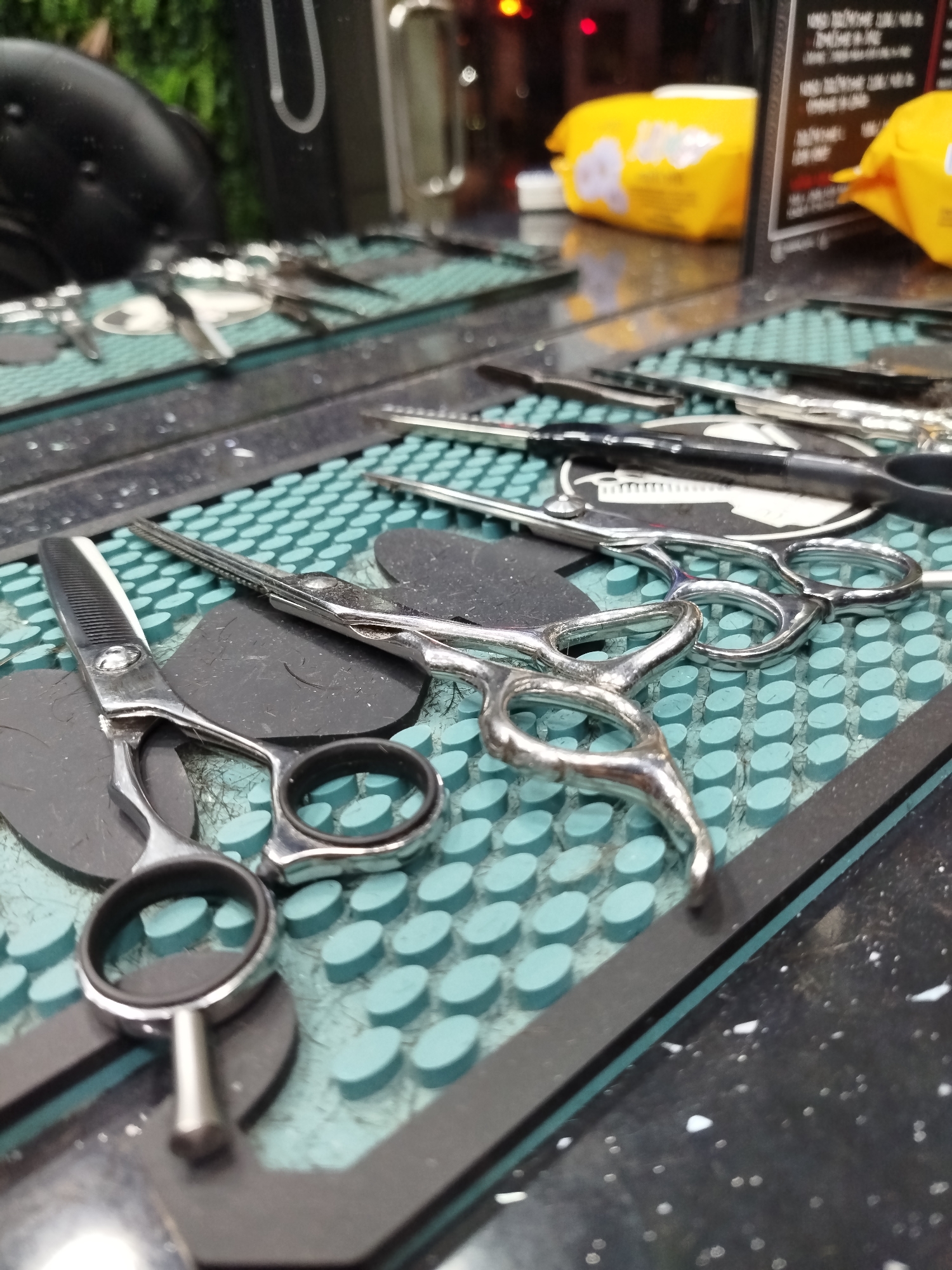 The image shows a close-up of barber tools, specifically various pairs of scissors, laid out on a teal colored mat with a grid of rounded bumps and a black border. The tools are mostly made of stainless steel, with some black accents. A scattering of cut hair can be seen beneath some of the tools. Other barber tools can be seen in the blurred background, along with a black barber chair, and yellow packs of wipes. The image appears to have been taken indoors at a barber shop. There is no clear evidence the scene is in Varna, Bulgaria.