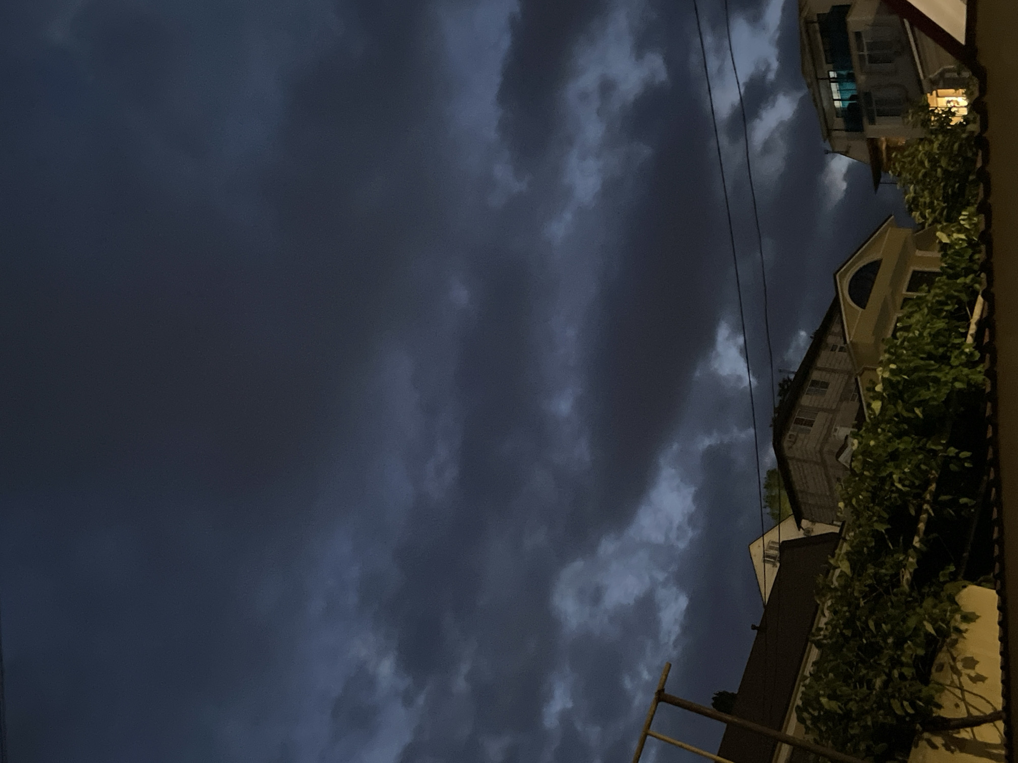 An overcast sky with dark, dramatic clouds looms over a cluster of buildings in Sochi, Russia. The image captures the tops of several houses, with visible architectural details such as pitched roofs, dormer windows, and balconies. Lush green foliage obscures the lower portions of the buildings, suggesting a temperate climate. Electrical wires are strung between the structures, indicating a developed urban area. The lighting is dim, characteristic of twilight or a heavily clouded day, with some artificial lights beginning to illuminate interiors of the buildings. The scene is static, with no visible people or ongoing activity.