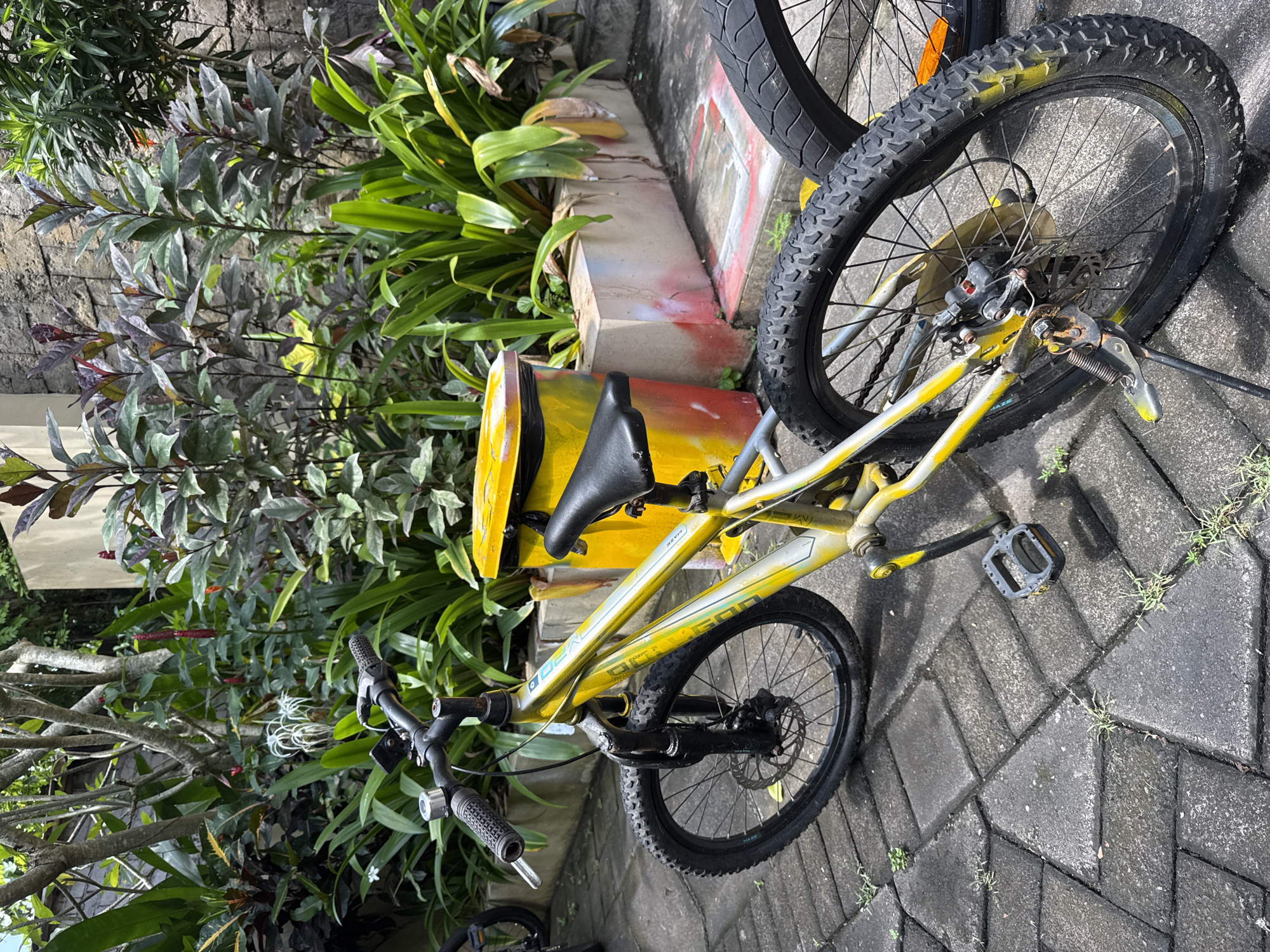 A yellow and silver mountain bike is parked on a paved surface, angled towards the viewer. The bike is positioned near a planter painted in yellow, orange, and red tones. Lush greenery surrounds the bike and planter, creating a backdrop of dense foliage. The scene is captured in daylight, suggesting an outdoor setting in Jimbaran, Indonesia.
