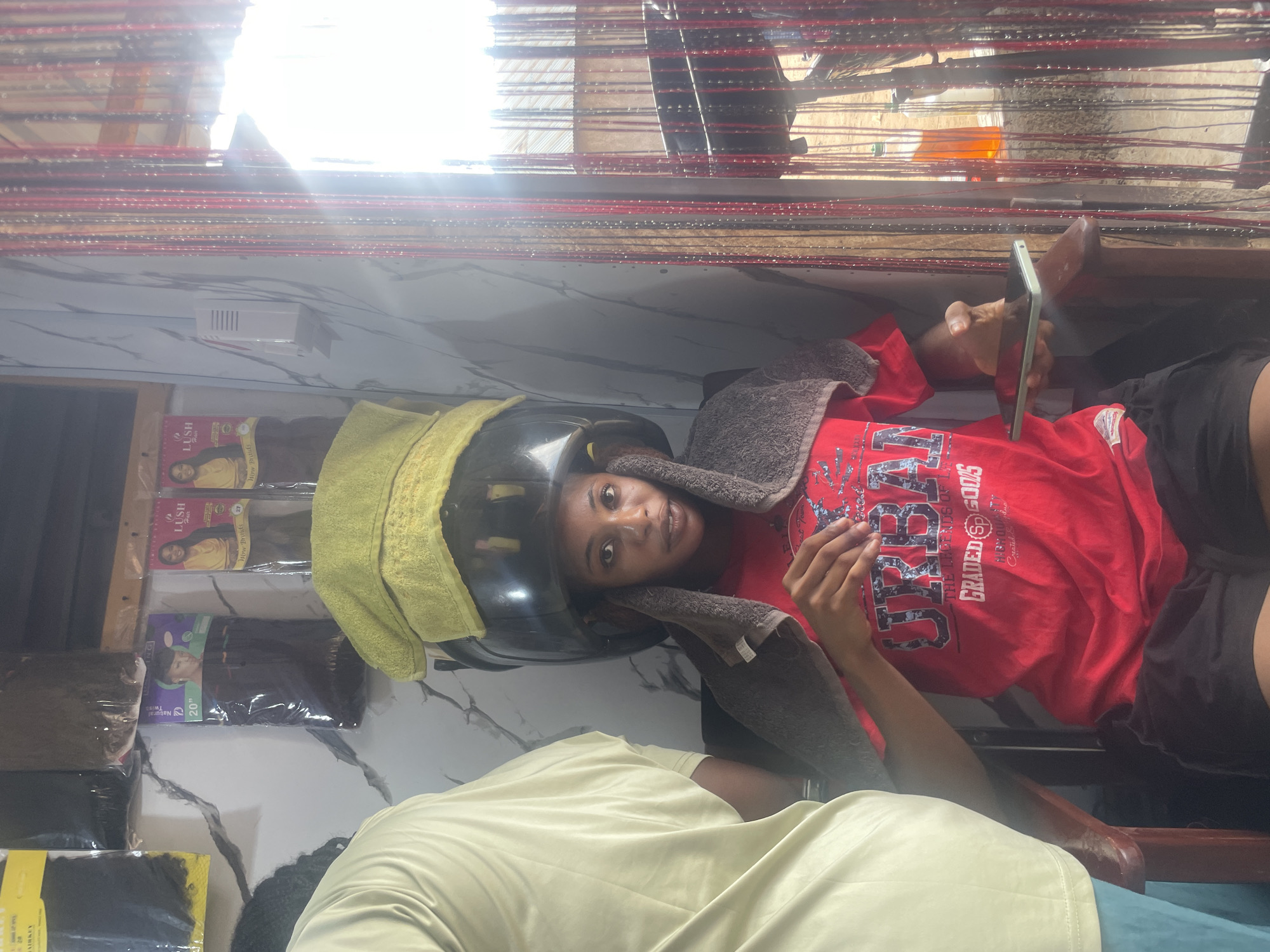 In Lafia, Nigeria, a young woman sits in a salon chair with a hair dryer helmet on her head, a yellow towel draped over it, and a grey towel around her neck. She is wearing a red t-shirt with "URBAN" and "GRADED GOODS" printed on it, and black shorts. Her right hand holds a smartphone. Beside her, another person, only partially visible from the back and side, wears a light yellow t-shirt and blue pants. The salon's interior features a wall with a marbled pattern, shelves displaying packages of hair extensions with branding such as "LUSH Hair," and a white wall-mounted device. A beaded curtain made of red and black strands hangs on the right side of the image, with a glimpse of an orange bottle and other items in the background. The scene is brightly lit, with sunlight streaming in from the right.
