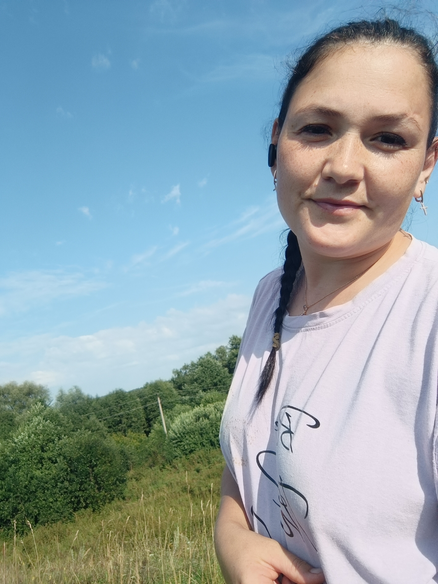 A selfie captures a woman with dark hair braided into a single plait, wearing a light purple t-shirt and a gold necklace. She has on hoop earrings, one with a cross charm. The background shows a clear blue sky with wispy clouds, a line of green trees, and tall grass. The location is not specified in the image.