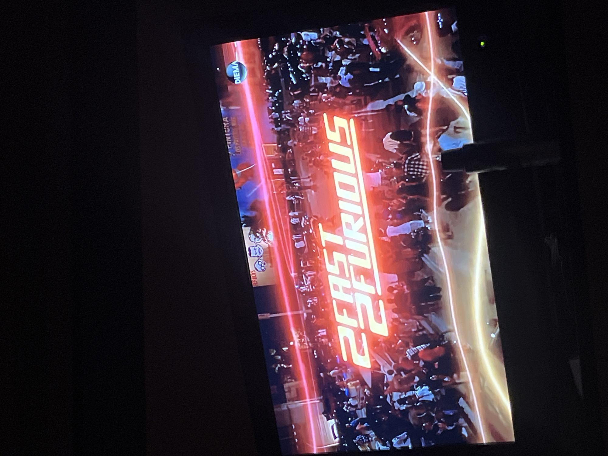 The image shows a television screen displaying the title card for the movie "2 Fast 2 Furious". The scene depicted on the screen appears to be a crowded outdoor event with numerous people present. A neon light or a laser line is projected across the scene.
The location of Veliko Tarnovo, Bulgaria is indicated by "FINIUKA LOS FASTS" displayed on the screen, referencing filming that occurred in that city. The logo "DIEMA" also appears, possibly referring to a Bulgarian television channel.