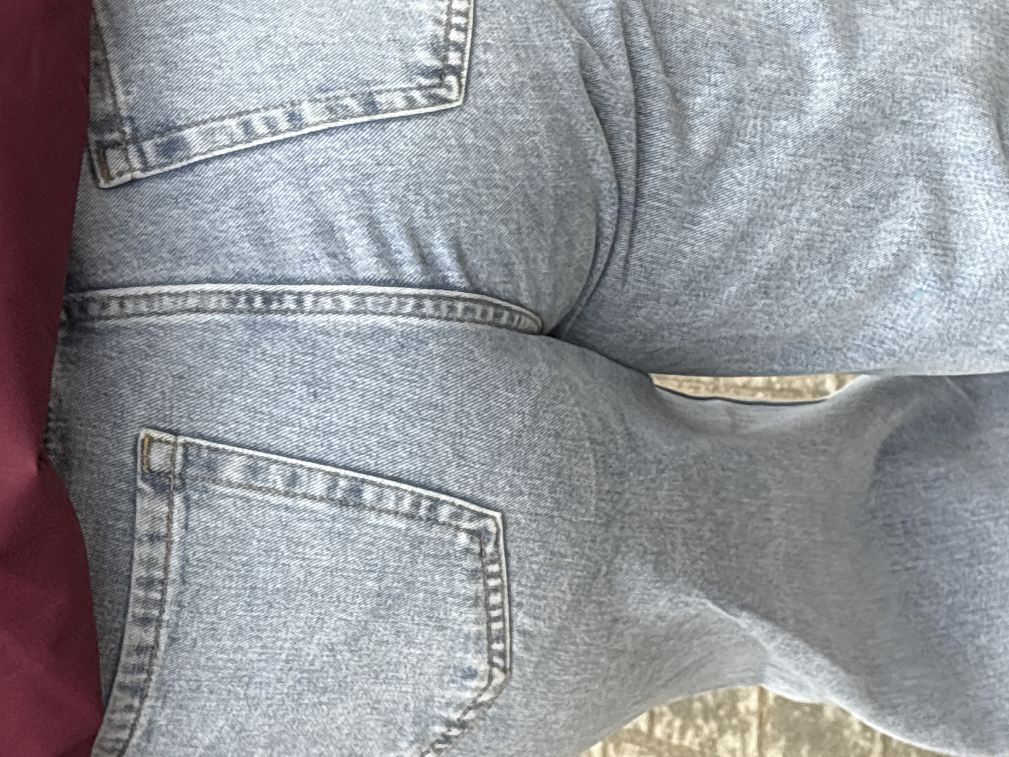 The image shows a close-up of light-wash denim jeans with visible back pockets and a red fabric partially visible on the left side. The denim is textured, and the stitching details of the pockets are prominent. The background appears to be a textured surface, potentially concrete or pavement. The location is not discernible from the visual information.