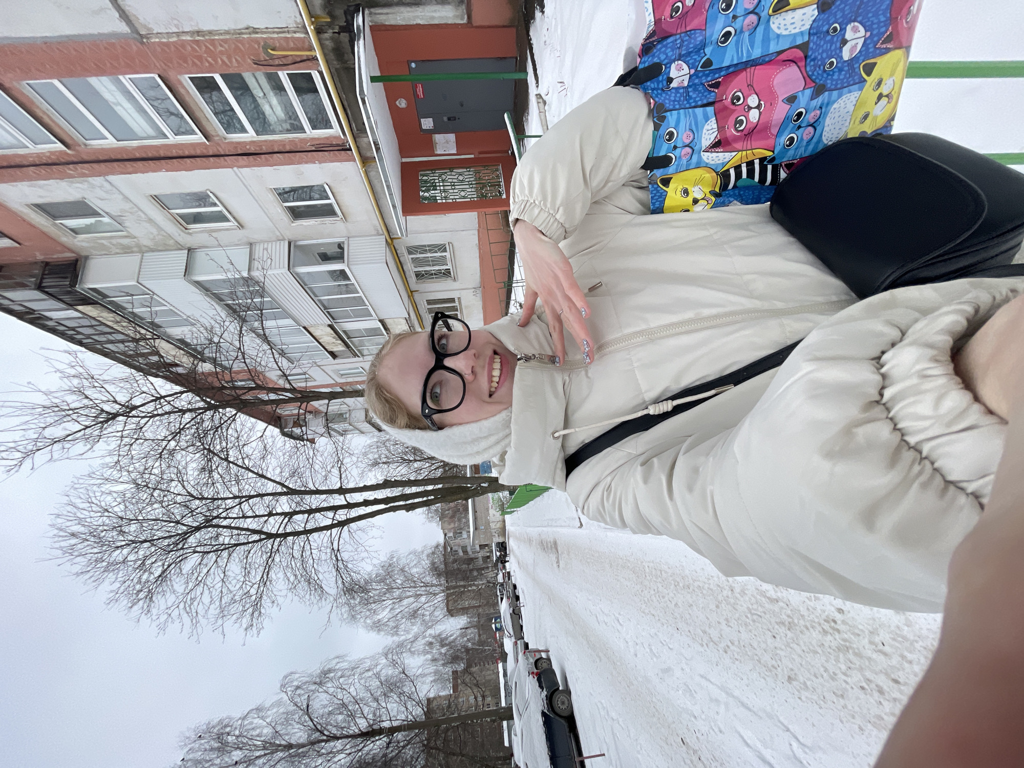 A young woman with eyeglasses is posing for a selfie in the snowy city of Izhevsk, Russia. She is wearing a white puffer jacket and carrying a colorful shopping bag depicting cartoon cats, as well as a small black purse. The background shows a typical urban street scene with apartment buildings, bare trees, parked cars covered in snow, and a snow-covered sidewalk.