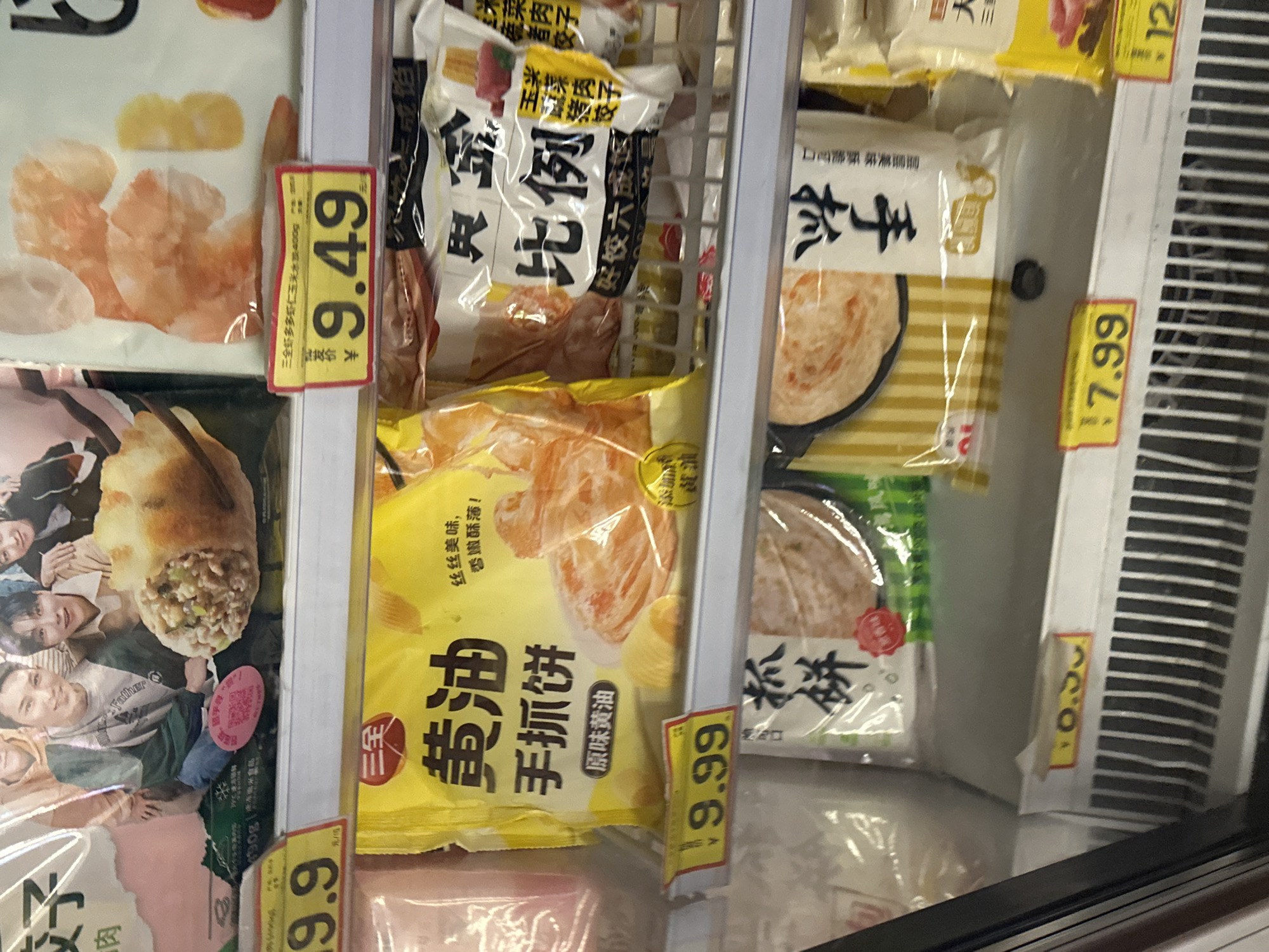 This image captures a close-up view of a refrigerated display case in a supermarket located in Chongqing High-tech Zone, China. The case contains multiple shelves stocked with various packaged frozen food items, primarily dumplings and pancakes, marked with price tags.
On the top shelf, a partially visible pink package on the left displays "¥ 9.9" on its price tag. To its right, a large package features a group of several young East Asian men and a prominent image of a fried, filled dumpling. A pink circular sticker with a QR code is affixed to this package. On the far right of the top shelf, a clear plastic bag contains what appears to be shrimp-shaped snacks. Beneath this item is a yellow price tag with a red border, indicating "¥ 9.49", and text above reading "三全多多虾仁玉米水饺400G".
The middle shelf features a bright yellow package on the left, labeled "黄油 手抓饼" (Butter Hand-grab Pancake) with "原味黄油" (Original Butter Flavor) and text indicating "丝丝美味, 酥脆诱人!" (Silky delicious, crispy and tempting!). It displays an image of cooked, layered pancakes. Below this package, a yellow price tag shows "¥ 9.99". To its right, a white and black package with prominent Chinese characters "比例" (Proportion/Ratio) is partially visible, featuring text "好饺六成饺", suggesting a type of dumpling, possibly corn and pork.
On the bottom shelf, a partially visible package on the left displays the characters "抓饼" (Hand-grab Pancake) and a red circular seal. The price tag below it is largely obscured. To the right, another package shows a cooked, layered pancake in a black pan, labeled "手抓饼" (Hand-grab Pancake). A yellow price tag below it clearly states "¥ 7.99". A further package is partially visible to the far right with a price tag of "¥ 12.XX" (partially obscured). The bottom of the freezer case features white horizontal grilles. No people are visible in the scene.