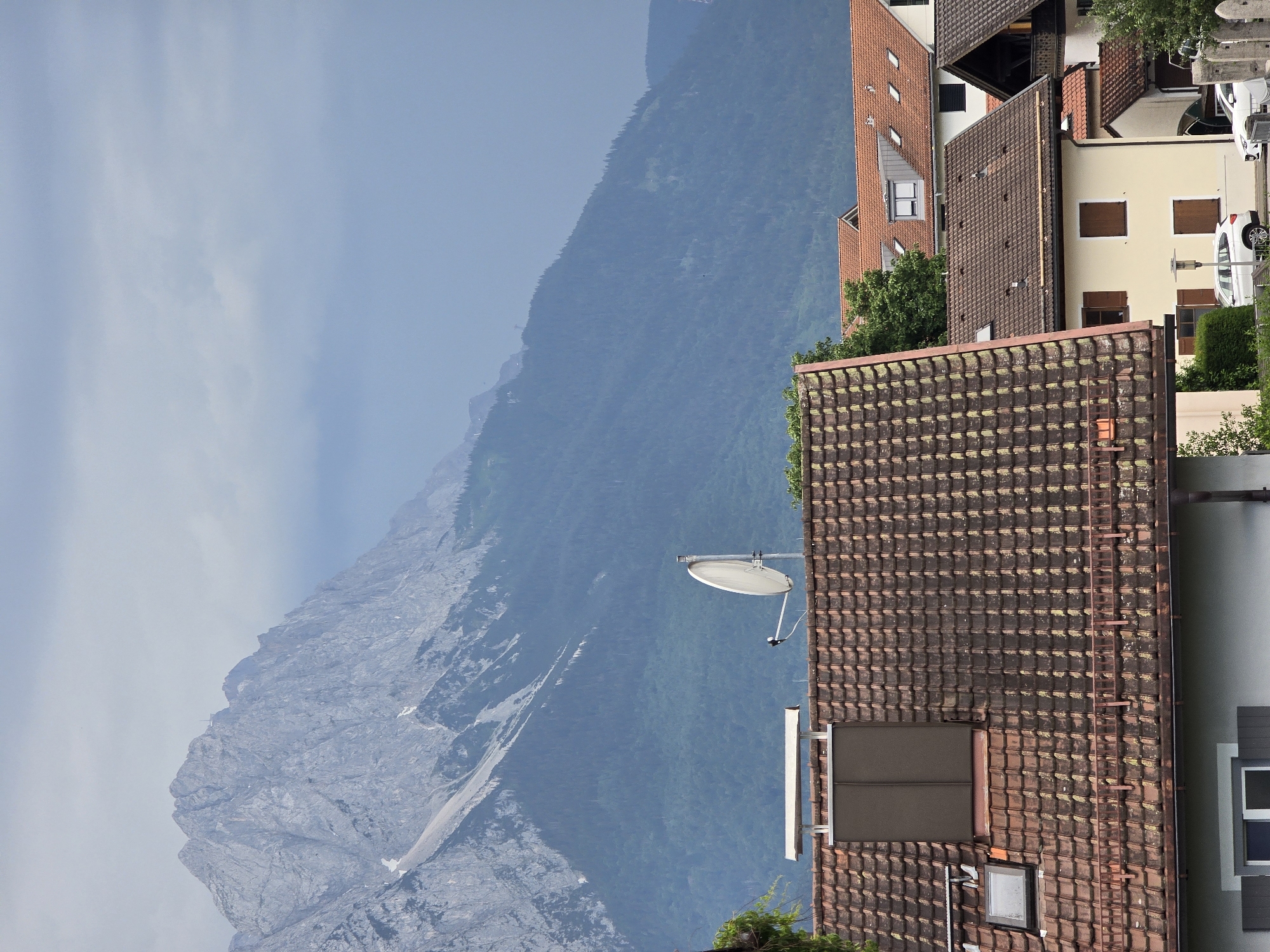 This is an outdoor, eye-level shot taken in a mountainous region, likely Garmisch-Partenkirchen, Germany. The image is vertically oriented. The foreground features the tiled roofs of several buildings, some appearing older with moss growth, while others are more modern. One building has a satellite dish mounted on its side. In the middle ground, a dense green forest covers a steep mountainside, giving way to a rugged, rocky mountain peak that dominates the background. The sky is a pale blue with some wispy clouds, suggesting a clear but possibly hazy day. The lighting indicates it is daytime, likely late morning or early afternoon. There are no people visible, but two white cars are parked on a street to the right of the buildings. The overall scene is serene and picturesque, showcasing a blend of residential architecture nestled against a dramatic natural landscape.