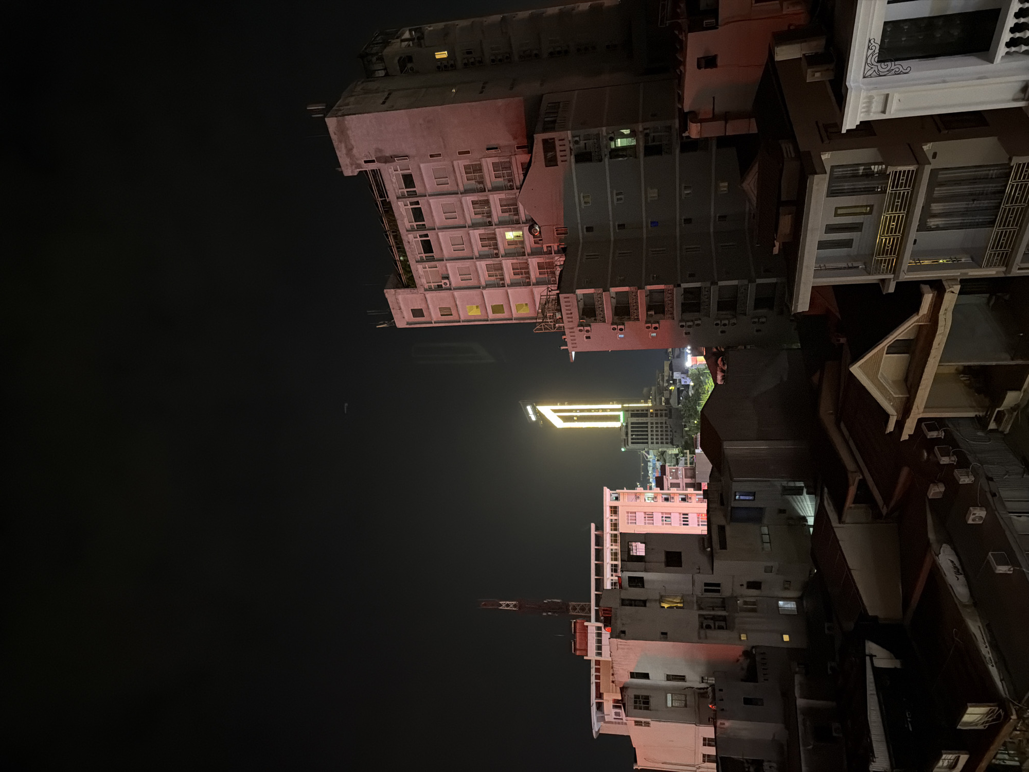 The image depicts a nighttime cityscape in Thuận Hóa Ward, Vietnam. The scene is dominated by multi-story buildings illuminated by artificial lights. In the background, a tall, slender skyscraper with a bright, neon-like yellow structure forms a prominent feature, suggesting it is a landmark. In the mid-ground, several apartment buildings or hotels are visible, with some windows lit, indicating occupancy. These buildings exhibit a mix of architectural styles and varying states of upkeep. In the foreground, a cluster of lower-rise buildings with dark roofs and visible air conditioning units are present, casting shadows and adding to the urban density. The sky is dark, with a faint hint of a distant airplane or satellite trail visible as a small white line. The overall impression is one of an active urban environment at night.