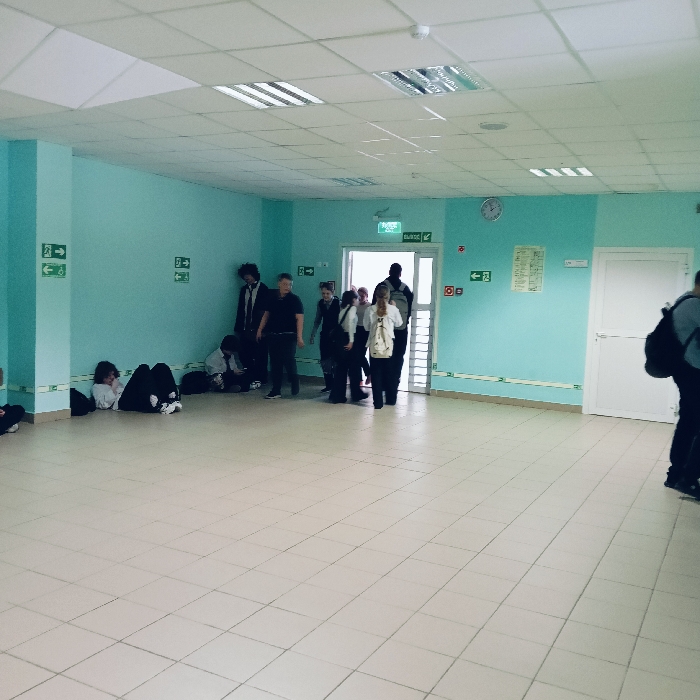 The scene depicts an interior corridor within a building in the city of Chaikovsky, Russia. The walls are painted a uniform light blue-green. The floor is covered with large, light-colored square tiles. The ceiling features a white acoustic panel grid system with recessed linear fluorescent light fixtures and a single smoke detector.
On the left side of the frame, two individuals are seated or lying on the floor against the wall. One person with dark curly hair and a white long-sleeved shirt is lying on their side. Another individual in dark clothing and white sneakers is seated with legs outstretched. A dark backpack is positioned on the floor near them. Standing near these individuals are three other people: one male with dark, curly hair wearing a dark suit jacket and tie; another male in a dark t-shirt and dark pants; and a third person partially obscured.
Towards the center-right, a group of approximately seven individuals is clustered around an open doorway. Above this doorway, a green sign with white text reads "ВЫХОД" (EXIT). This group appears to be in motion, either entering or exiting the corridor. A large male in a dark shirt is prominent within this group. A female wearing a white top and dark pants, carrying a light-colored shoulder bag, is also visible.
Multiple green and white emergency exit signs with directional arrows are mounted on the walls, pointing both left and right. A circular wall clock with a white face and dark numbers is visible on the right wall, along with a rectangular document or poster and two red square signs. Further right, a closed white door with a handle is visible. On the far right, a person with a dark backpack is partially visible, moving away from the camera. The corridor is well-lit by overhead artificial lighting.