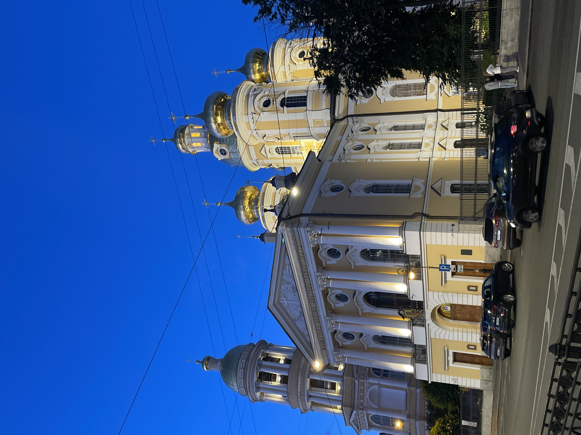 The image shows a view of the Smolny Cathedral in Saint Petersburg, Russia, during twilight. The cathedral's yellow facade and gilded domes are illuminated by artificial lights, contrasting with the deep blue of the evening sky. Power lines are visible crisscrossing the sky above the building. In the foreground, a street is lined with parked cars and a black car is in motion on the right side of the frame. A few individuals are standing on the sidewalk near a fence. The overall scene depicts an urban environment at dusk, with the prominent religious structure serving as the central subject.