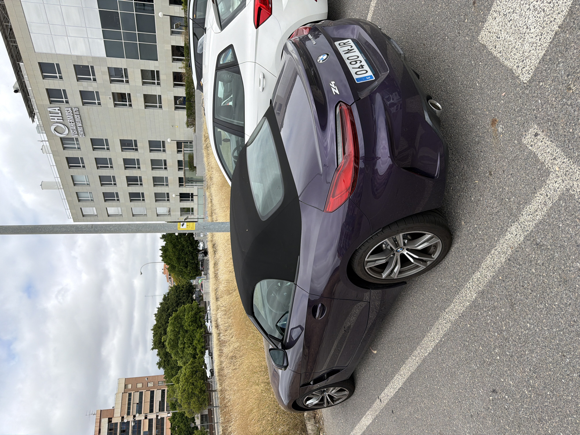 A purple BMW Z4 convertible is parked in a lot next to a white SEAT Leon. The BMW has its black soft top up. The SEAT is a five-door hatchback. In the background, a modern, light-colored building with a grid of windows displays the sign "HLA VISTAHÉRMOSA CENTRO MÉDICO V76." The building is situated behind a strip of dry, tall grass and a row of green trees. Several other cars and a street lamp are visible in the distance. The sky is overcast.