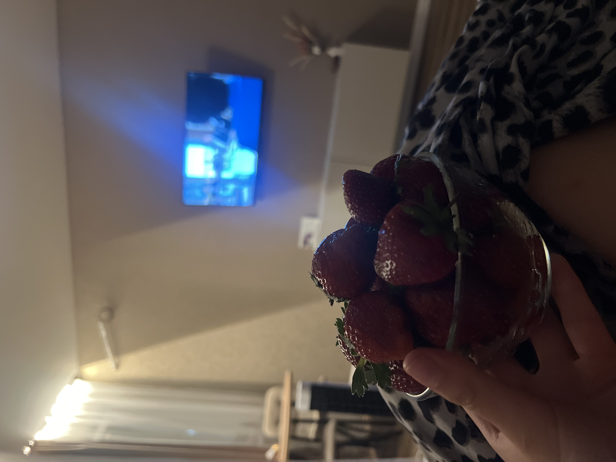 A close-up, indoor shot shows a person holding a glass bowl filled with fresh strawberries. The strawberries are a deep red color and appear ripe. In the background, a television screen displays a bright blue image, and a wall-mounted cabinet is visible below it. A curtain hangs on the left side of the frame, and a table with chairs can be seen through it. The scene is illuminated by soft indoor lighting, casting shadows on the wall. The overall impression is one of a cozy evening at home. This image does not contain information that could be used to determine the location in the city of Krasnodar, Russia.