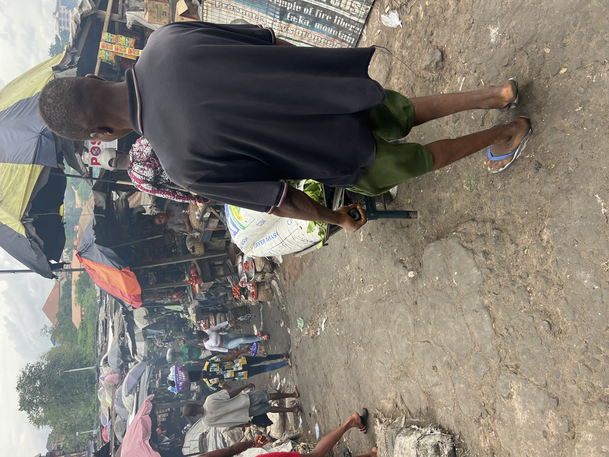Here is a detailed description of the image:
**Location:** The image depicts a crowded street market in Nkpor, Nigeria.
**Environment:** The ground is rough, uneven, and appears to be worn asphalt or concrete. The market is congested with makeshift stalls covered by tarps and umbrellas for shade. There is evidence of cooking and burning, with smoke visible in the background. Buildings and vegetation are visible in the distance.
**People:**
* A man in a black shirt, green shorts, and blue flip-flops is pulling a wheelbarrow. The wheelbarrow contains a large white sack.
* Various other people are walking through the market, including women in patterned clothing, a person in a white shirt and shorts, and individuals whose faces are obscured by the crowd or angle of the photo.
**Objects:**
* Wheelbarrow with a white sack.
* Market stalls selling food items.
* Umbrellas and tarps providing shade.
* Signage with text related to nutrition and religious references visible in the upper part of the frame.
* Various items for sale including food, vegetables, baskets, and other merchandise.
**Actions and Interactions:** People are walking and shopping in the market. The man in the black shirt is pulling the wheelbarrow through the crowded street.