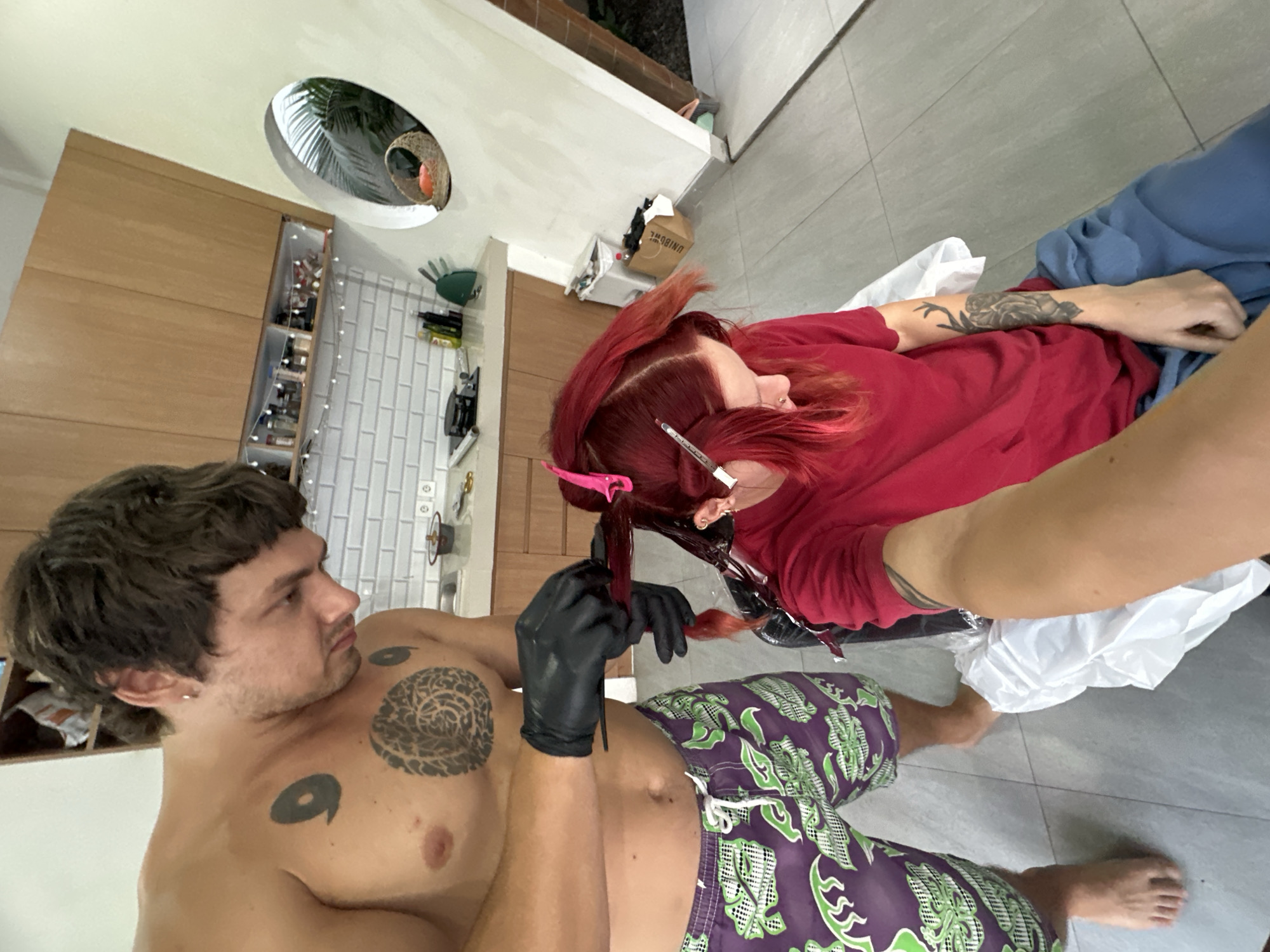 The image shows a couple in what appears to be a home setting in North Kuta, Indonesia, likely a bathroom or a well-lit indoor space with tiled flooring. The person in the foreground, a man with dark, tousled hair and multiple tattoos including a large circular design on his chest and a smaller one on his torso, is shirtless and wearing purple and green patterned shorts. He is looking to his right, off-camera.
The second person, a woman with vibrant red hair, is seated and partially visible in the upper right portion of the frame. She is wearing a red top and blue pants, and has tattoos on her arm. She is actively applying hair dye to her own hair using black gloves and a comb. Pink hair clips are used to section her hair. The scene suggests a DIY hair dyeing session.
In the background, there are wooden cabinets with shelves stocked with various items, suggesting a storage area. A window or skylight with greenery visible through it is also present on the ceiling, hinting at a tropical environment. A string of fairy lights is draped along the shelves, adding a decorative element. There are also some tools and supplies on a wall rack, consistent with a grooming or creative space. The overall mood is casual and intimate, capturing a personal moment between the couple. No specific time of day or weather is discernible, but the lighting suggests it could be daytime.
