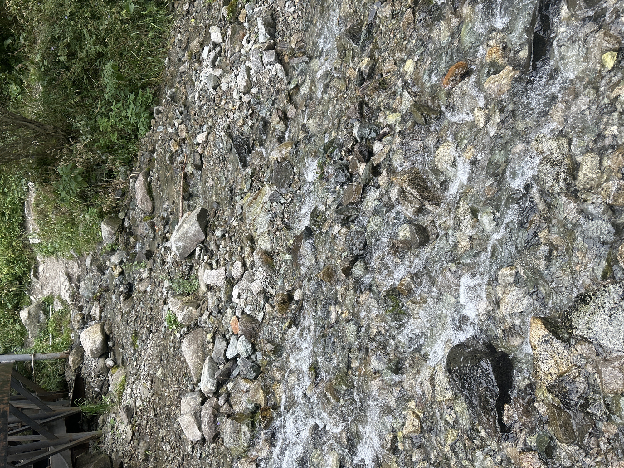 The image shows a rocky stream flowing over a bed of various sized stones. Lush green vegetation grows on the bank to the right and in the background, where a path is also visible. To the left, the dark, wooden structure of a bridge or walkway is partially visible. The water is clear, revealing the stones beneath, and there are some white water rapids in the foreground. The scene appears to be in a natural outdoor environment, possibly in a mountainous or park area. This specific image does not contain enough visual information to definitively place it within the city of Almaty, Kazakhstan.