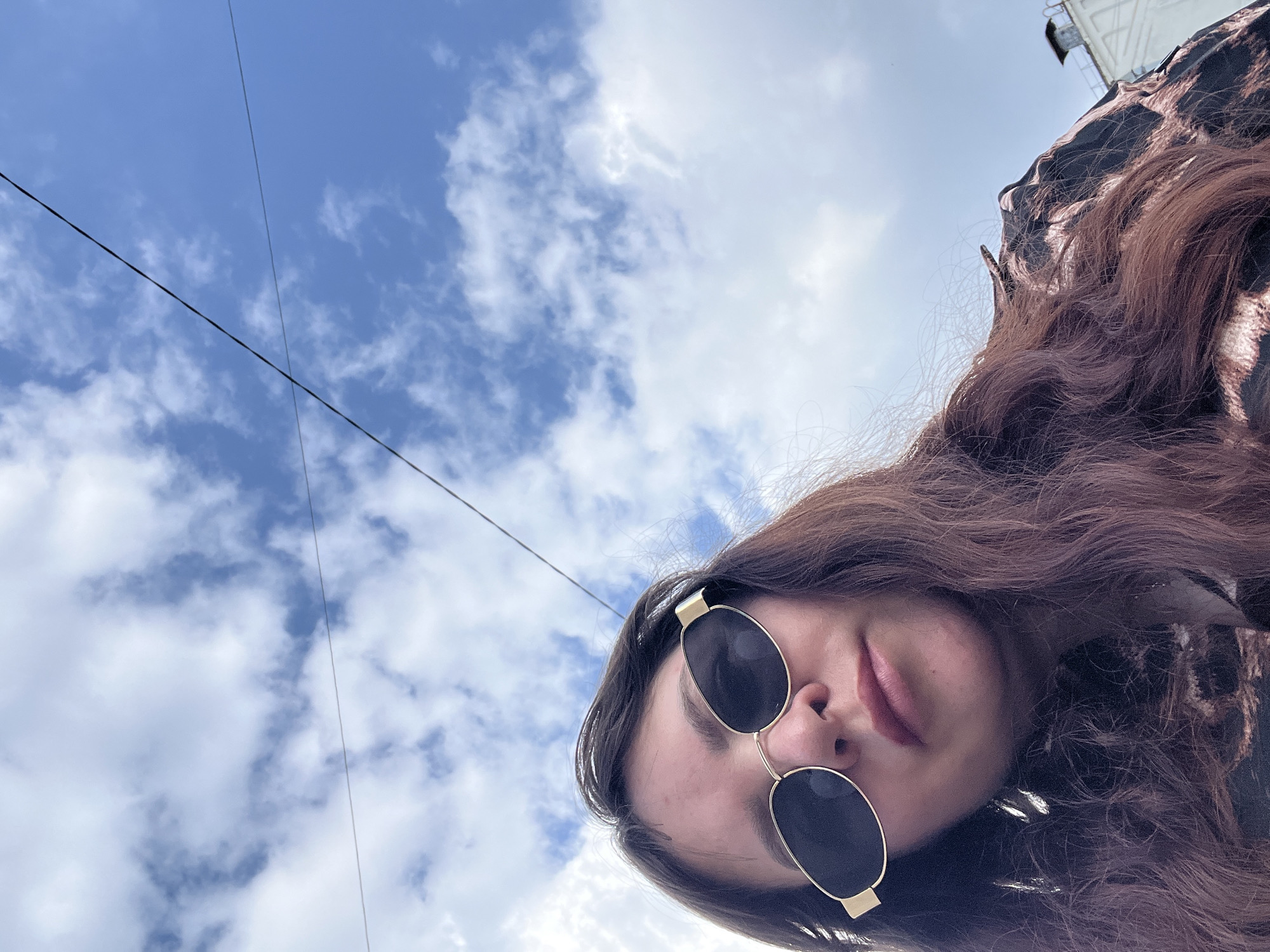 The image shows a close-up, low-angle selfie of a woman wearing oval sunglasses and a patterned shirt. She has long, wavy brown hair. The background is a bright blue sky with scattered white clouds, and a thin wire is visible overhead, suggesting an urban environment. Based on the context provided, the location is Noviy, Russia. The lighting suggests it is daytime. There are no other people or discernible objects in the immediate vicinity.