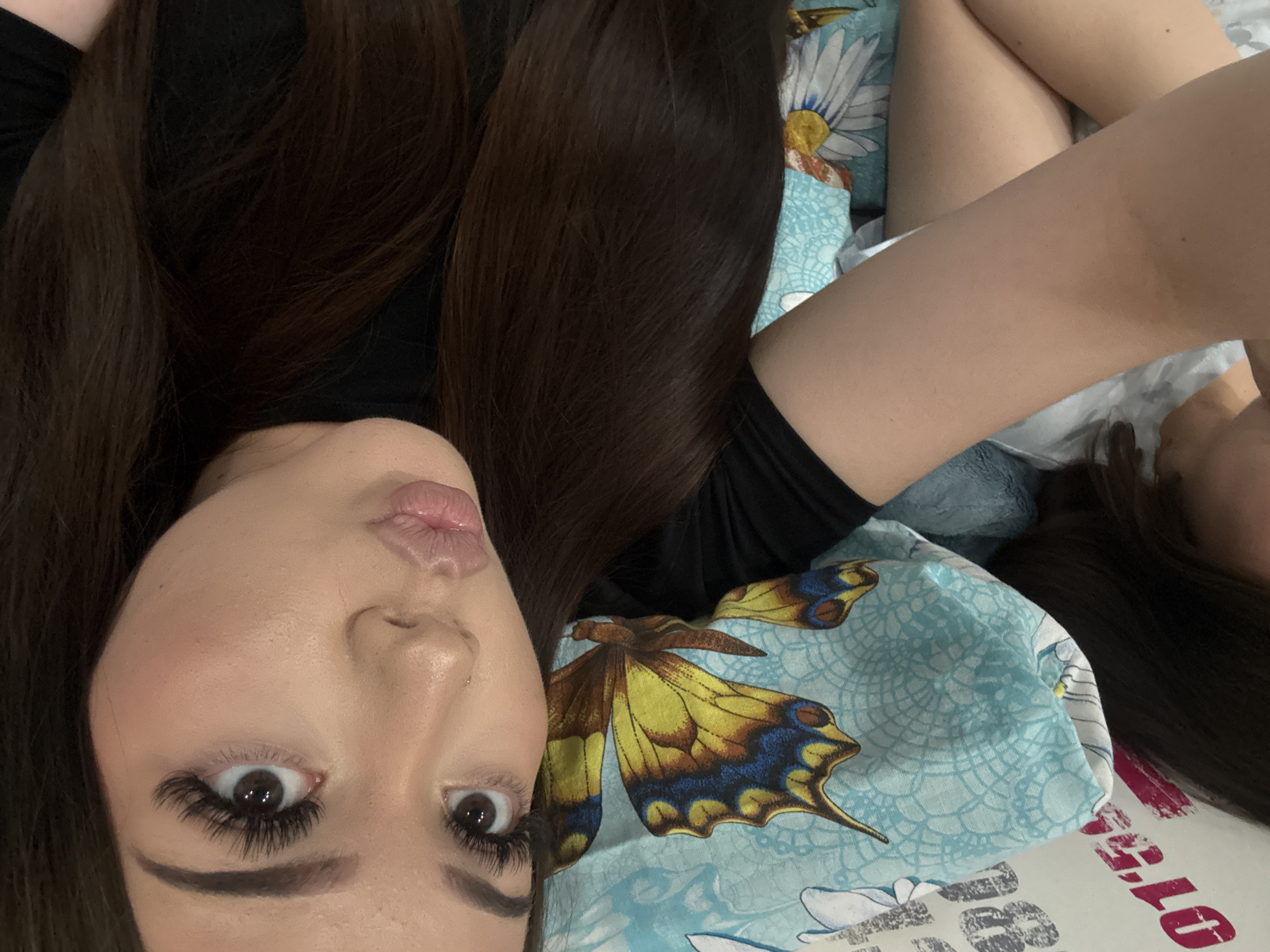 The image shows a close-up, overhead shot of a young woman lying down, with her head tilted and her hair spread out on a pillow and sheets. The woman is wearing a black top. Her makeup includes dramatic false eyelashes and lip gloss. The pillow and sheets have a blue background with a pattern of yellow and blue butterflies and white daisies. On the lower left side of the image, there are white and pink numbers, possibly part of a fabric or poster. The overall scene appears to be indoors, in a bedroom or living space. No specific location in Naberezhnye Chelny, Russia is identifiable from the visual information provided.