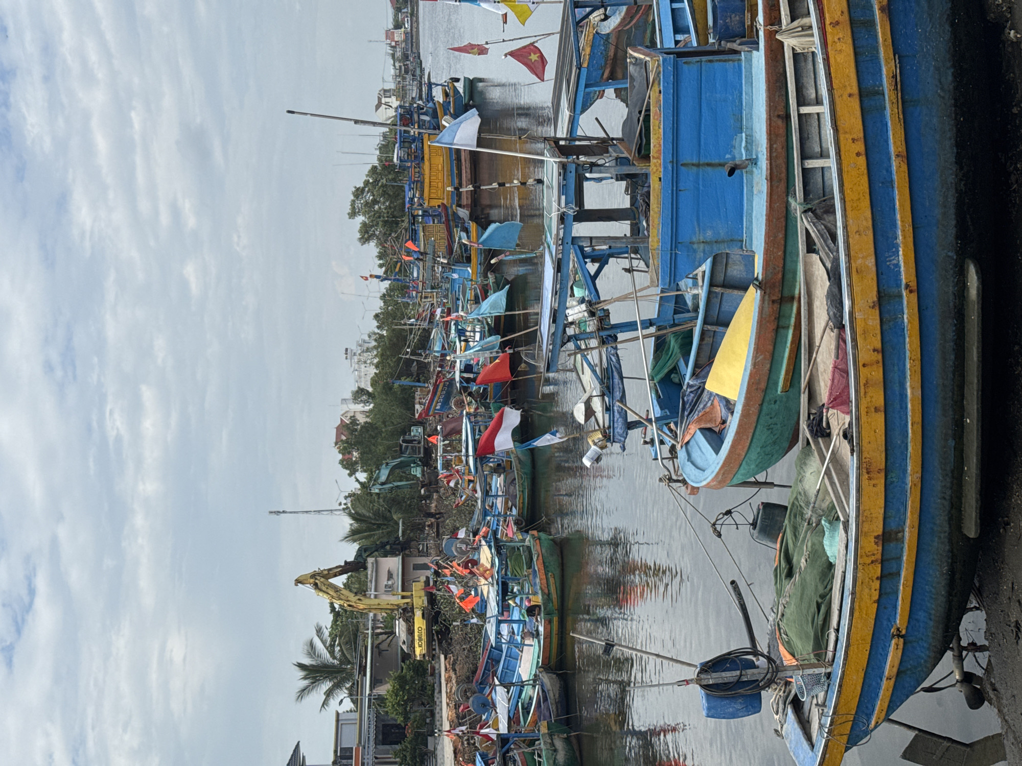 The image depicts a busy harbor in Phú Thủy, Vietnam, filled with numerous fishing boats of various sizes and colors. The boats are moored closely together, creating a dense network on the water. Many of the vessels are adorned with flags, some national flags and others smaller, colorful flags, indicating a likely maritime or fishing activity. In the background, construction equipment, including excavators, is visible, suggesting ongoing development or dredging work along the waterfront. Buildings and trees line the shore, with a glimpse of distant structures and a tall communication tower. The sky is overcast, with a mix of clouds and lighter patches, indicating a diffused daylight. The overall scene conveys a sense of vibrant, industrial, and maritime activity within a coastal town.