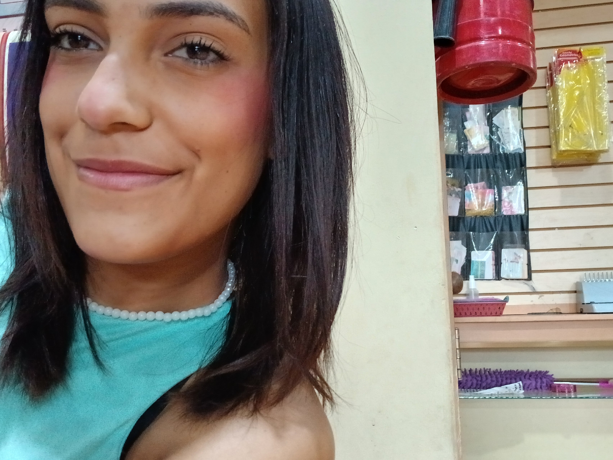 The image shows a close-up, medium shot of a young woman in what appears to be a store or market setting, possibly in Juan Griego, Venezuela. She is looking towards the camera with a slight smile. Her face is well-lit, highlighting her brown eyes, defined eyebrows, and rosy blush on her cheeks. She has dark, straight hair that falls around her shoulders. She is wearing a turquoise-colored top and a white beaded necklace.
In the background, to the right of the woman, there are shelves displaying various items. These include what appear to be packages of stationery or craft supplies, a red bucket or container, and possibly some cleaning supplies. The shelves are made of wood and are mounted on a light-colored wall. The overall environment suggests a retail or service-oriented establishment. The lighting in the scene is moderately bright.