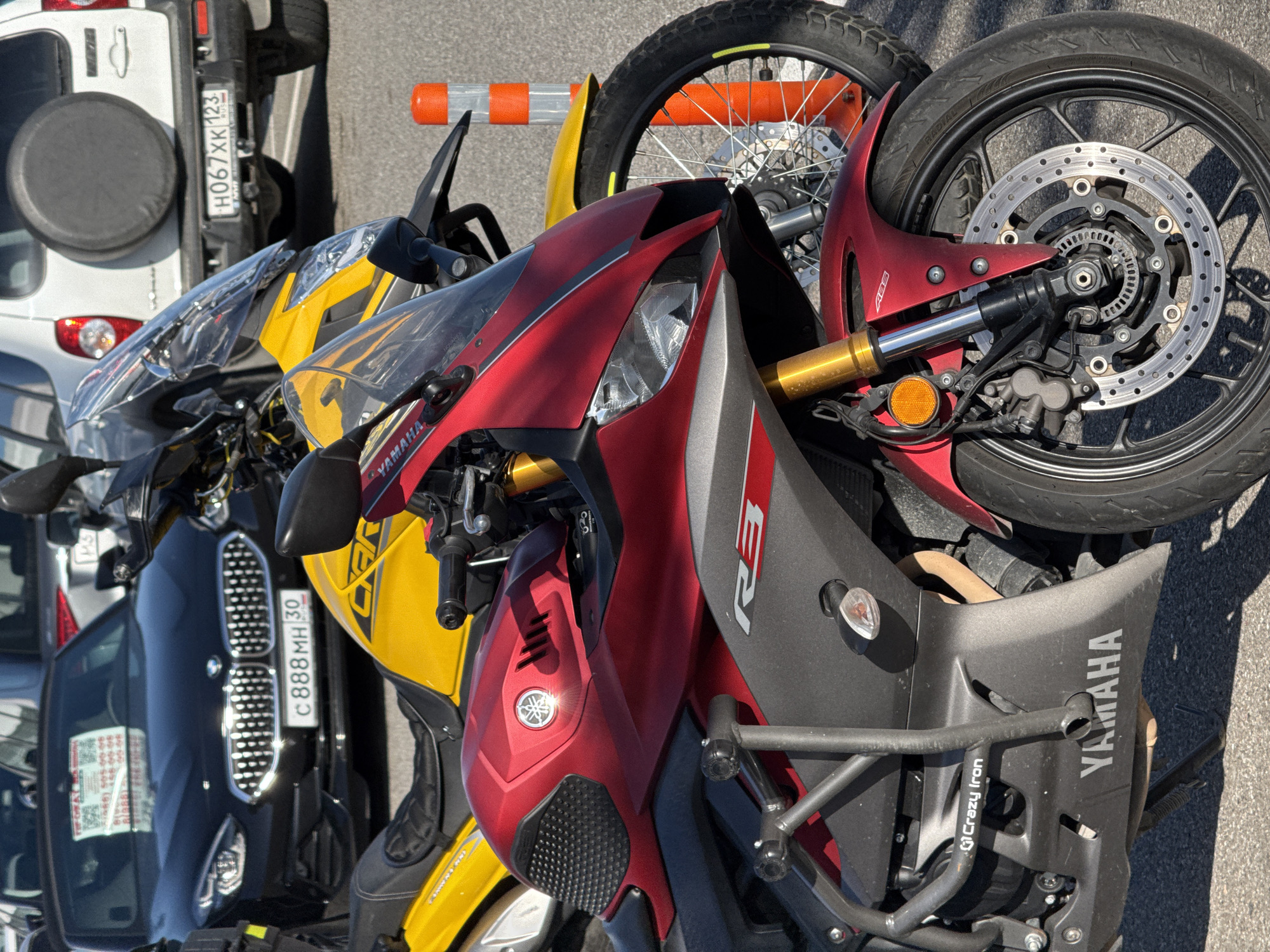 The image shows a parking area in the city of Sirius, Russia. Three vehicles are visible: a white SUV with license plate number H067XK 123, a black BMW with license plate number C888MH 30, and two motorcycles, a red Yamaha R3 and a yellow motorcycle next to the black BMW. The image focuses on the red Yamaha R3, with its front wheel prominently displayed. The Yamaha R3 is equipped with crash bars. An orange and white traffic barrier is also visible, lying on the ground, near the yellow motorcycle. The scene appears to be a typical parking area in an urban setting.