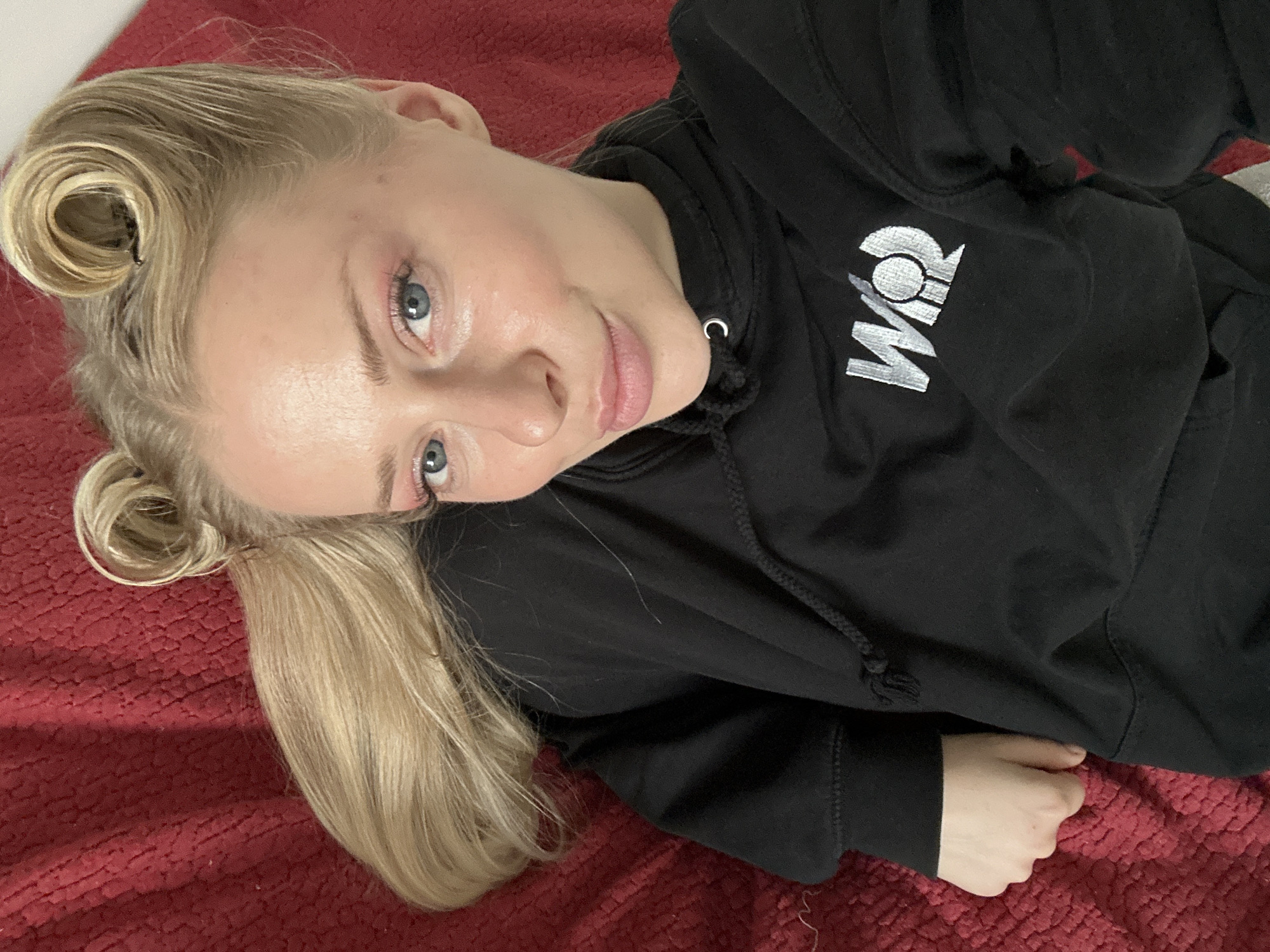 The image shows a young woman with blonde hair styled in two buns on the top of her head, lying on a textured red surface. She is wearing a black hoodie with a white logo on the left chest. Her eyes are open and she is looking towards the camera. The lighting suggests an indoor setting. There is no information in the image to suggest a location in Krakow, Poland.
