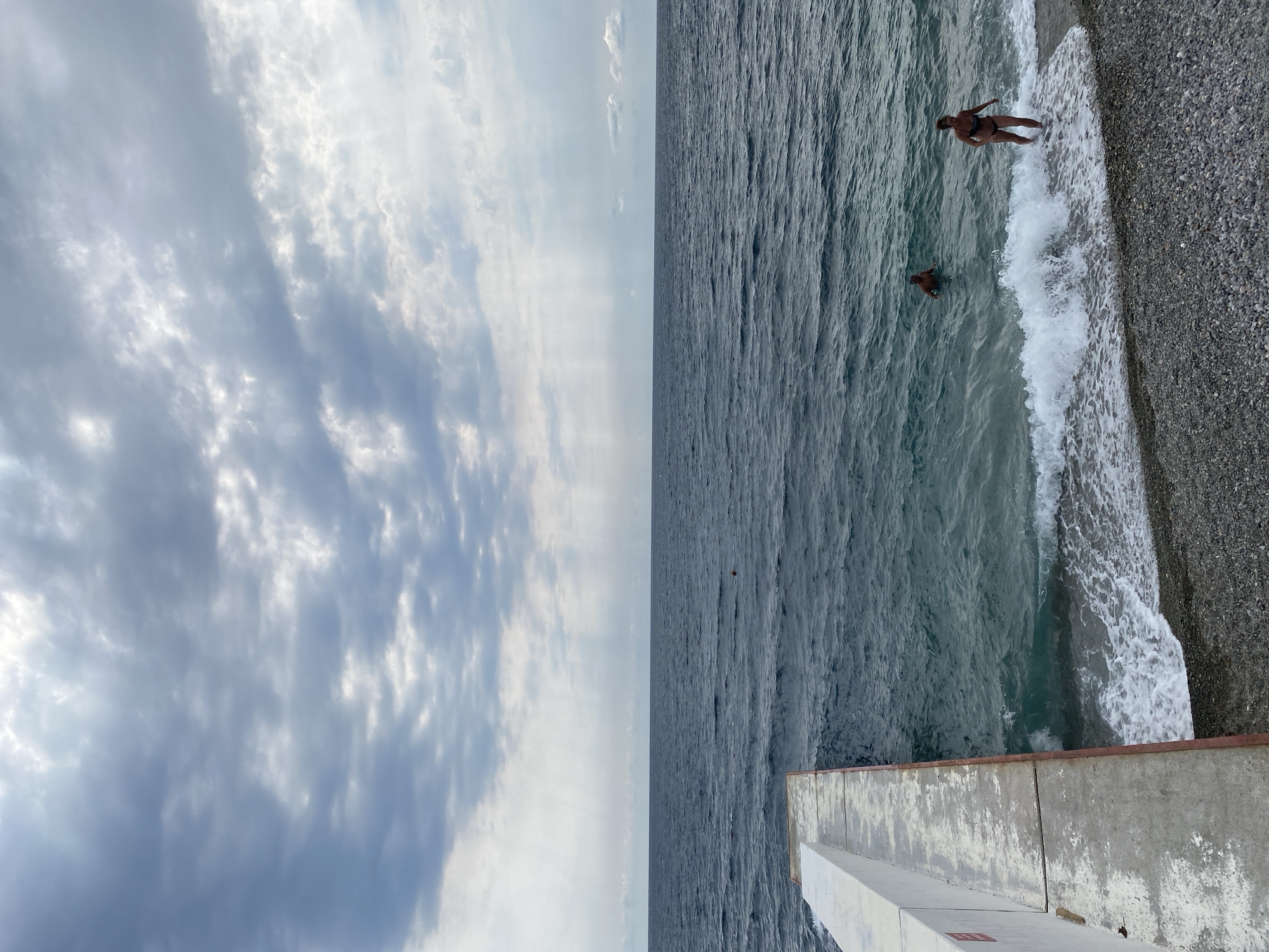 The image depicts a beach scene with two individuals in the water and a concrete pier. The sky is overcast with dramatic clouds, and shafts of light break through in the distance. The sea is choppy, with waves reaching the pebble-strewn shore. A woman in a dark bikini is walking out of the water, with a wave breaking around her. Further out in the sea, a man is swimming. The pier, made of concrete with a red trim, extends from the left side of the frame into the water. The location is identified as Gasprynskyi settlement council, Ukraine.
