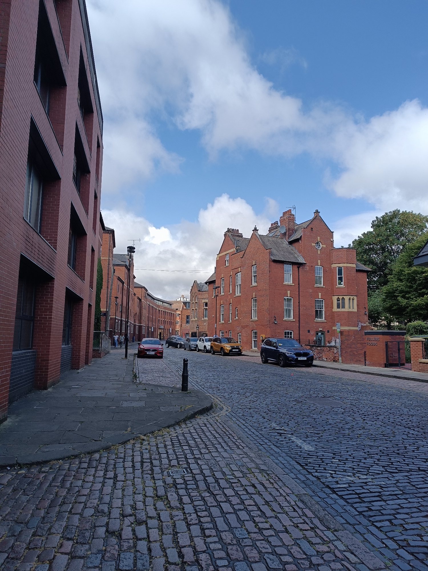 The image depicts a cobblestone street in Leeds, United Kingdom, lined with red brick buildings. The street curves to the right as it recedes into the distance. On the left, a modern red brick building with dark-framed windows dominates the foreground. The right side of the street features older brick architecture, including a prominent building with a turret and a sign that reads "FULLERS FOODS". Several cars are parked along the street, including a red hatchback, a yellow SUV, and a dark blue SUV. The sky is blue with scattered white clouds, and trees are visible in the background on the right. A few pedestrians are walking further down the street on the left. The scene appears to be a quiet urban environment on a daytime.