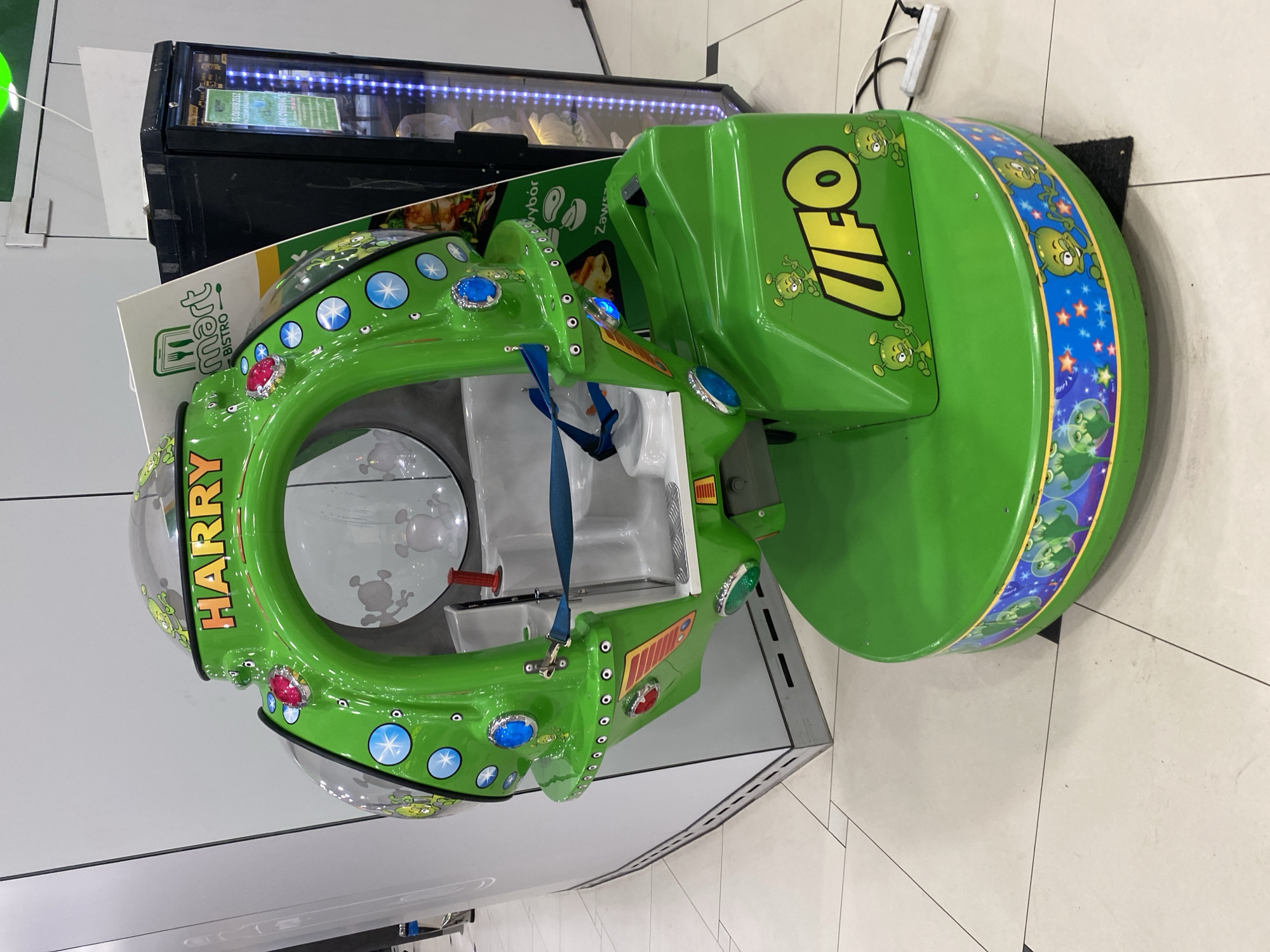 The image shows a close-up, eye-level shot of a brightly colored, green children's amusement ride, shaped like a flying saucer with an opening for a child to sit inside. The ride is decorated with cartoon aliens and stars, and prominently features the word "UFO" in yellow lettering on its base. A sign that reads "HARRY BISTRO" is visible on the ride's upper section. To the right of the ride is a tall, black refrigerated display case with a glass door, illuminated by blue LED lights, suggesting it is part of a commercial establishment such as a convenience store or a food service area. The floor is tiled in a light beige color with dark grout lines, indicating an indoor setting, possibly a shopping mall or a play area. A white power strip with a black cord is plugged into an outlet on the floor near the ride. The overall scene is clean and well-lit. The object itself is a kiddie ride located at a "smart bistro" in Szczecin, Poland.
