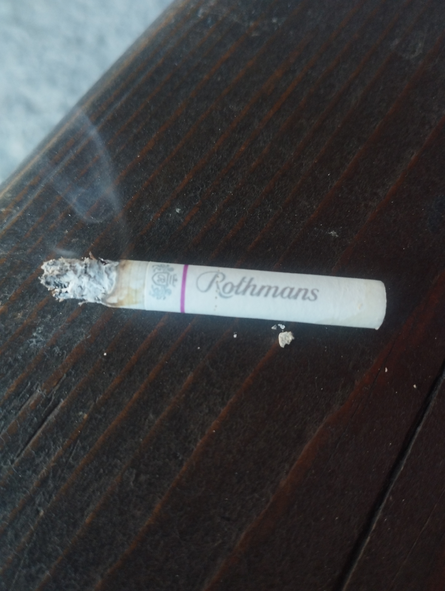 The image displays a close-up, eye-level shot of a lit cigarette with the brand name "Rothmans" visible. The cigarette is positioned horizontally on a dark, wooden surface with visible grain. Ash has accumulated at the tip of the cigarette, and wisps of smoke are rising from it. There are a few small, light-colored particles scattered on the wooden surface near the cigarette, possibly ash or debris. The background is blurred, showing a lighter, textured surface. No specific location within the city of Homyel, Belarus, can be identified from the provided image.